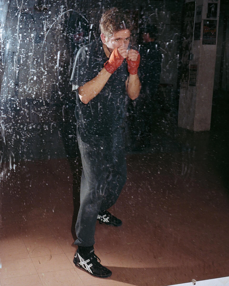 King, Queen, Knave by Gregory Halpern, photographed by Gregory Halpern — A man in dark sweats and red hand wraps holds a boxing guard, seen through a sheet of clear plastic flecked with paint.