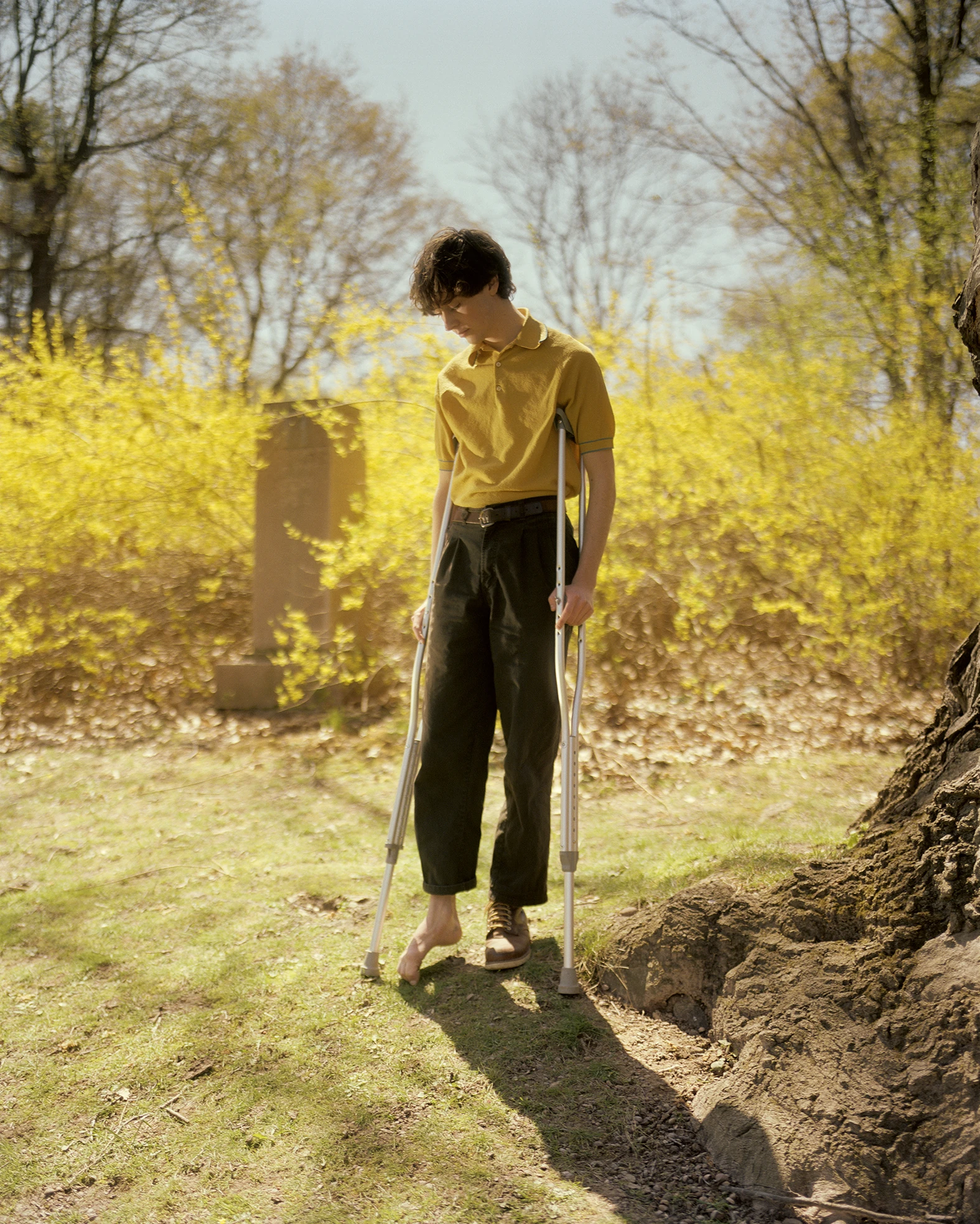 King, Queen, Knave by Gregory Halpern, photographed by Gregory Halpern — A young man on aluminium crutches, one foot bare and one in a boot, stands on grass between a headstone and a wall of yellow forsythia in flower.
