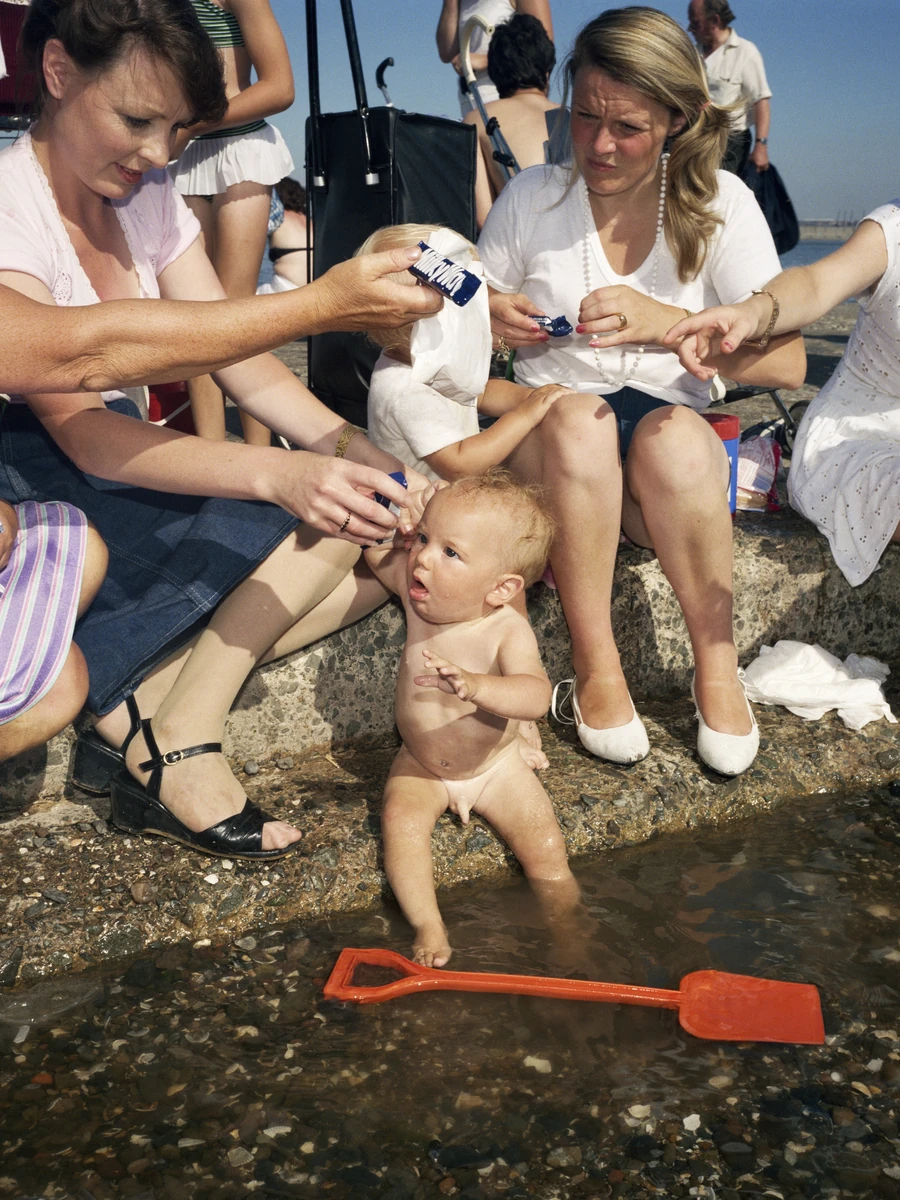 The Last Resort by Martin Parr, photographed by Martin Parr — A naked baby stands in a shallow pool of water at the edge of a concrete slipway, four women in summer dresses leaning down from the stone step with ice creams and lolly wrappers, a red…