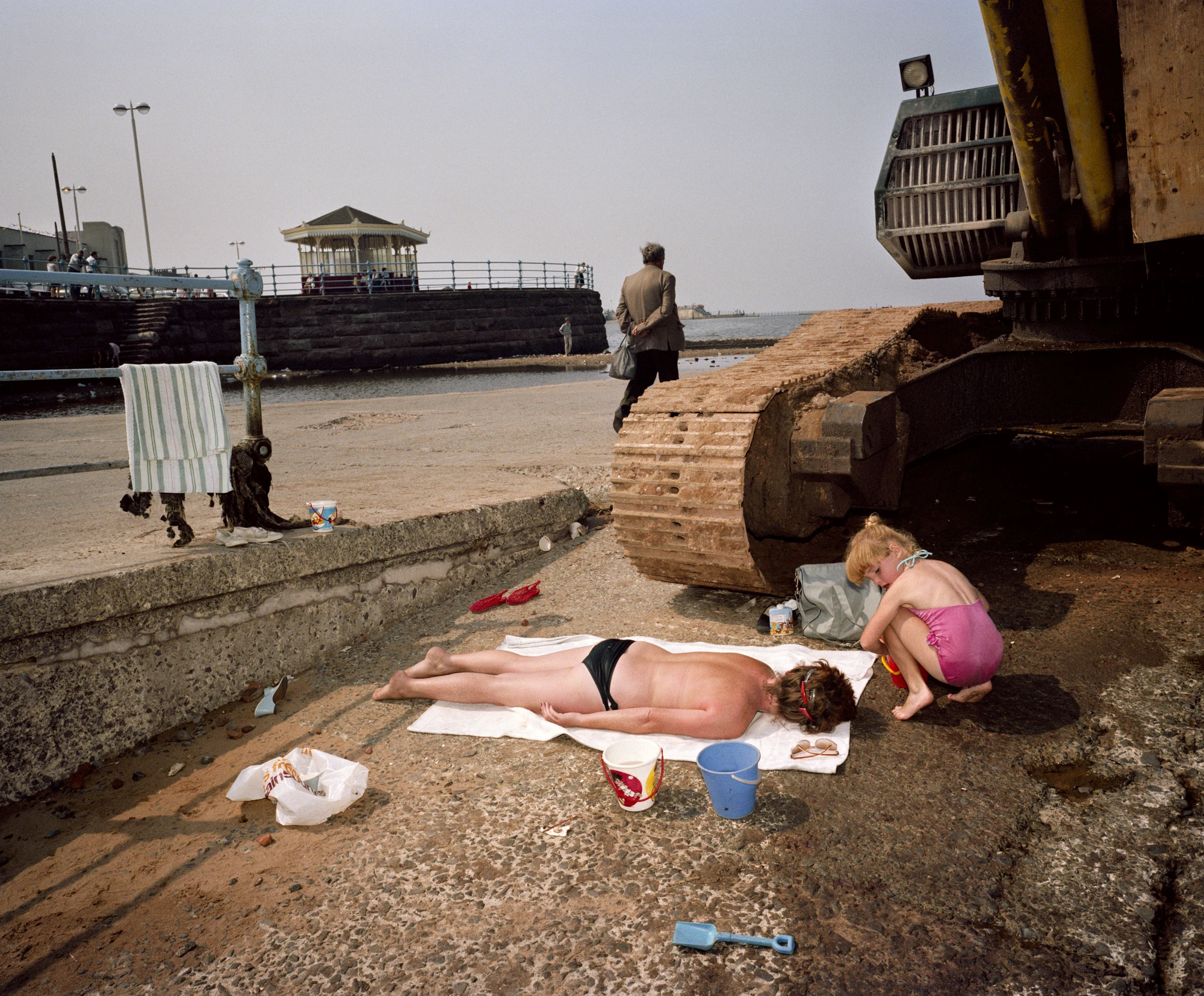 The Last Resort by Martin Parr, photographed by Martin Parr — A woman sunbathes face down on a white towel laid over cracked concrete beside the rusted caterpillar track of an excavator, a girl in a pink swimsuit crouching in its shadow, a bandstand and…