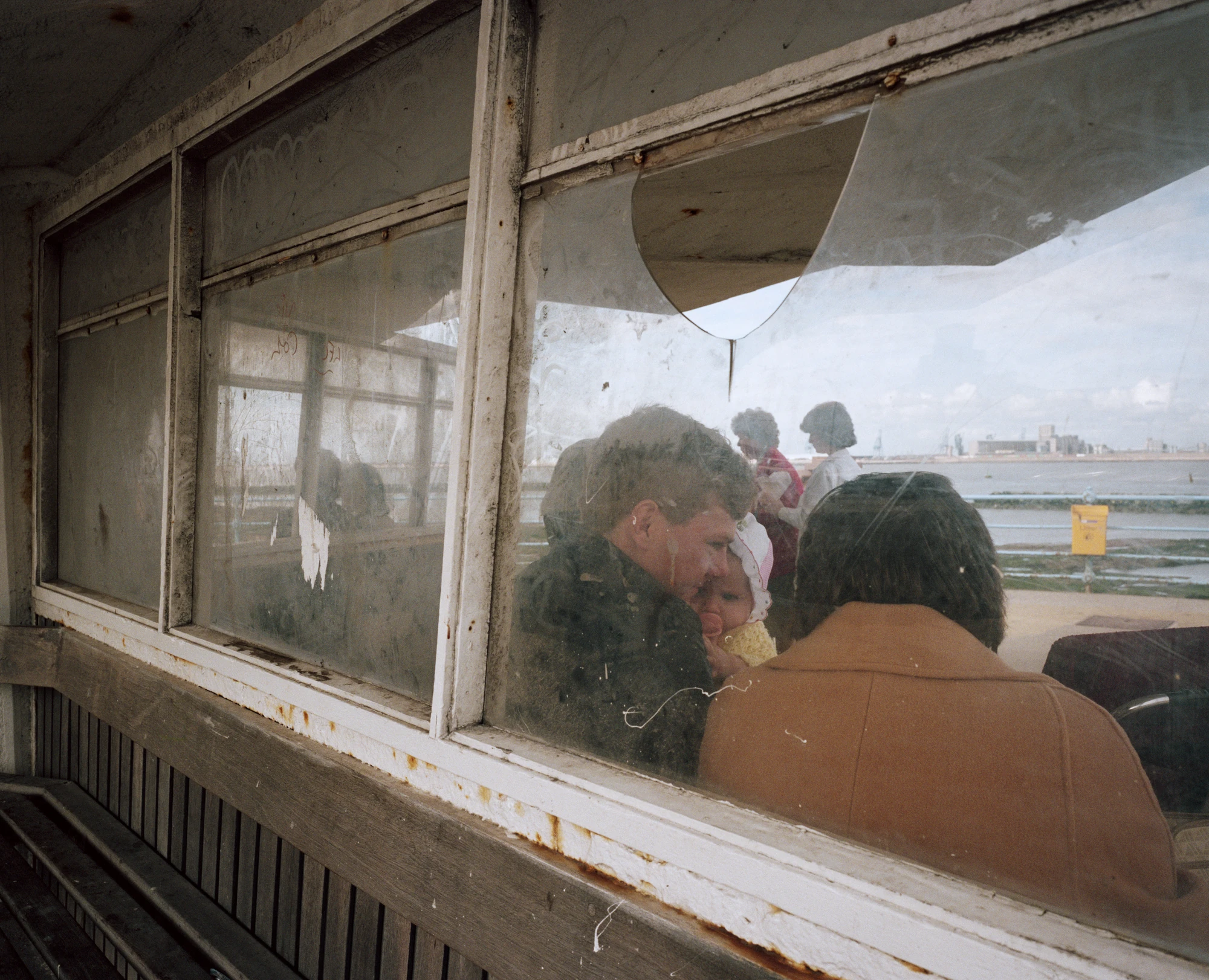 The Last Resort by Martin Parr, photographed by Martin Parr — Through the scratched and broken glass of a promenade shelter, a woman in a dark coat holds a baby in a bonnet, the pale estuary and dock cranes visible past the peeling white window frame.
