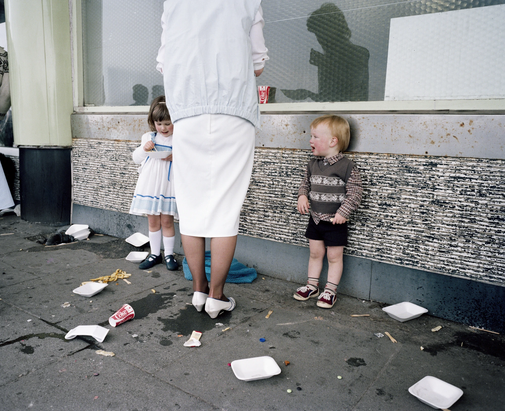 The Last Resort by Martin Parr, photographed by Martin Parr — A woman in a white skirt and heels stands at a serving hatch with her back to the camera, a small girl eating from a tray at her left, a red haired toddler bawling against the wall at her…