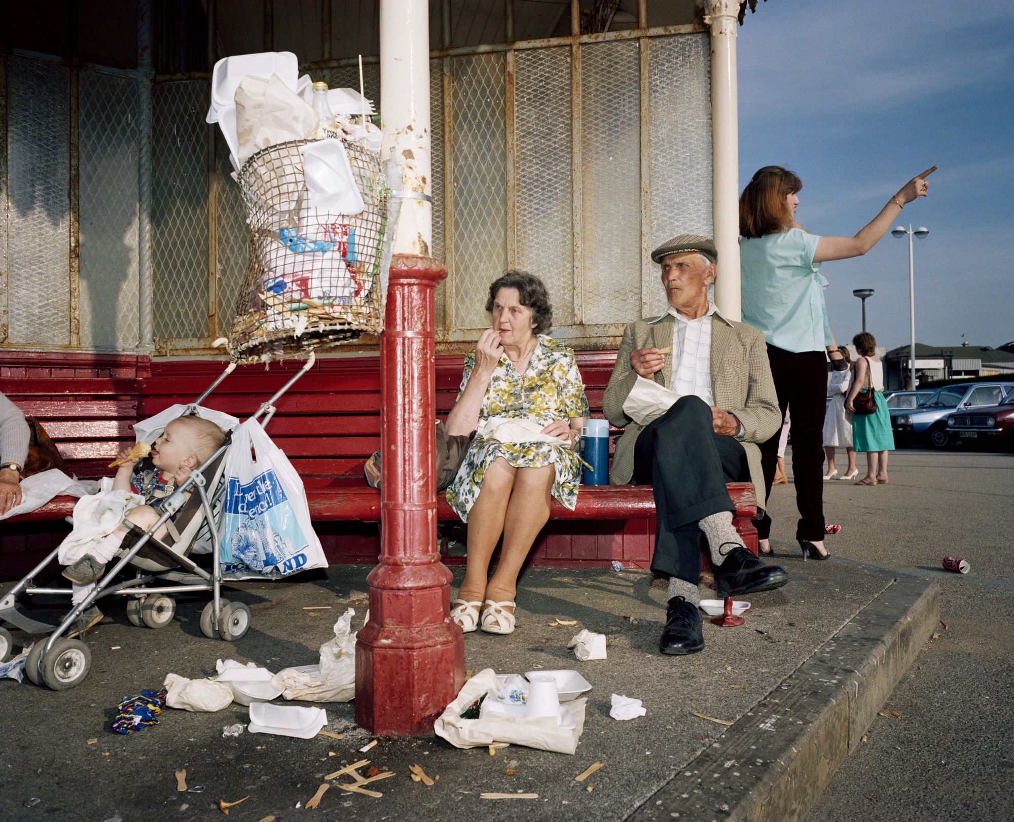 The Last Resort by Martin Parr, photographed by Martin Parr — An elderly couple eat chips from paper on a red painted bench under a wire litter bin overflowing with polystyrene trays, a baby asleep in a pushchair with an ice cream, wooden forks and…