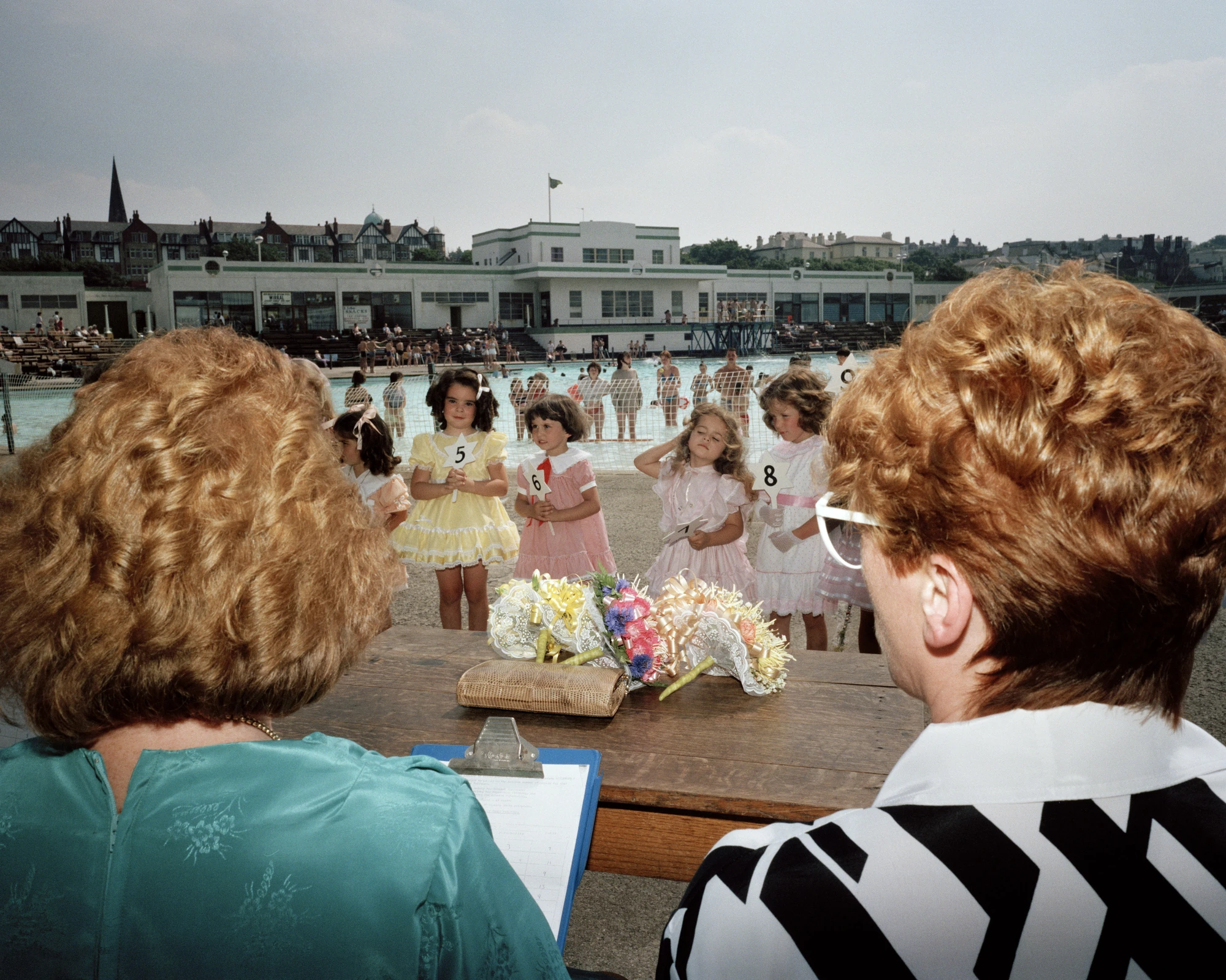 The Last Resort by Martin Parr, photographed by Martin Parr — Seen over the shoulders of two judges at a wooden table, small girls in yellow and pink frilled dresses hold numbered cards beside the lido pool, the white deco pavilion and terraced houses…