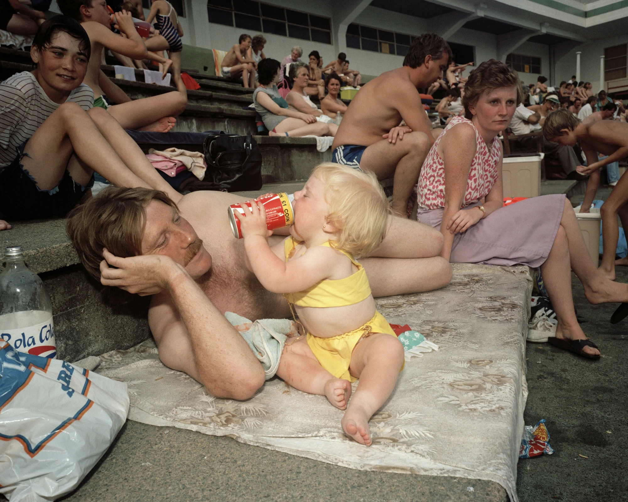 The Last Resort by Martin Parr, photographed by Martin Parr — A man lies back on a patterned towel spread over the lido's concrete terracing while a toddler in a yellow bikini drinks from a can propped against his chest, bathers packed along the tiers…