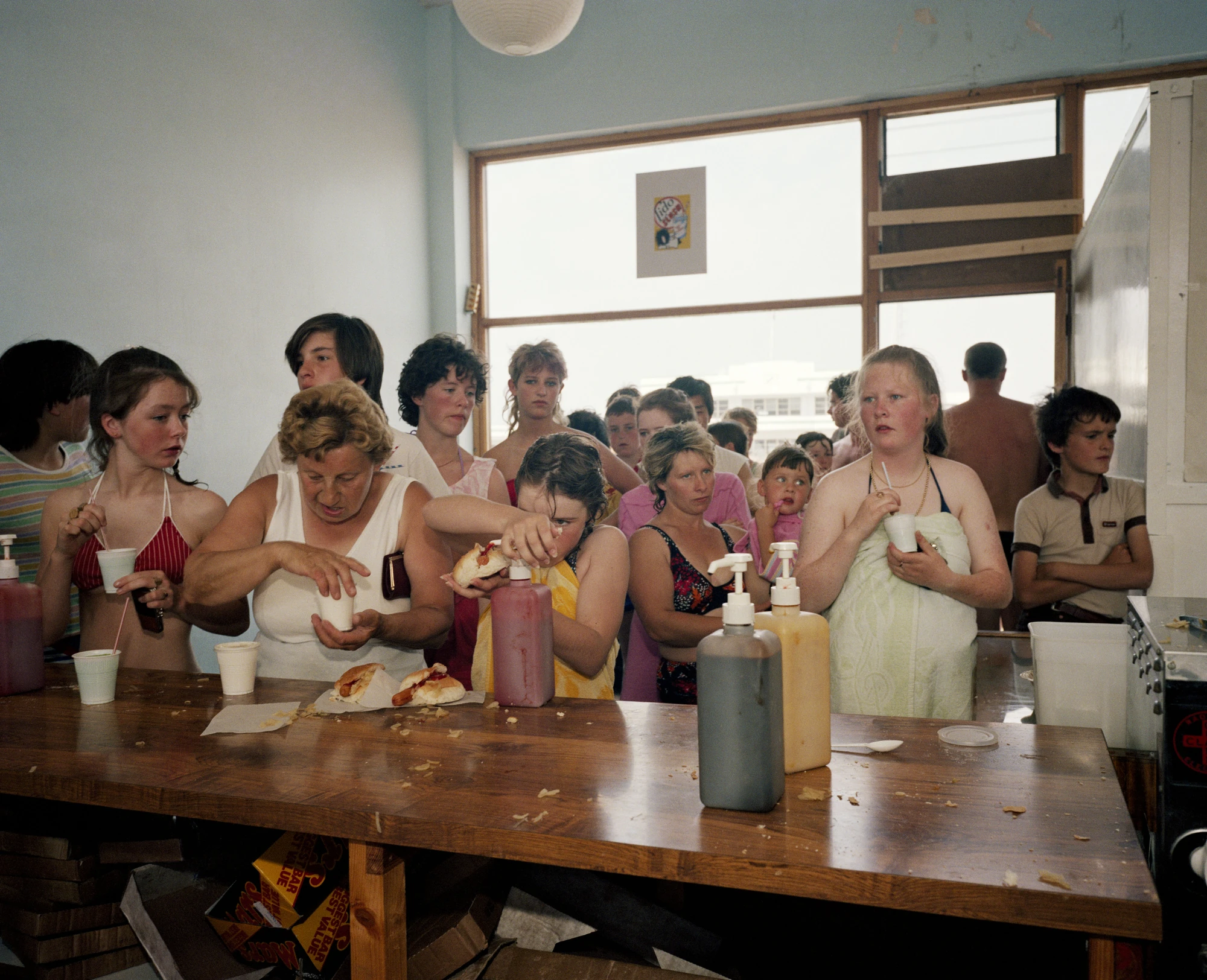 The Last Resort by Martin Parr, photographed by Martin Parr — A crowd in swimsuits and towels presses along a wooden snack bar counter loaded with hot dogs, ketchup and mustard dispensers and plastic cups, a poster taped to the window behind.