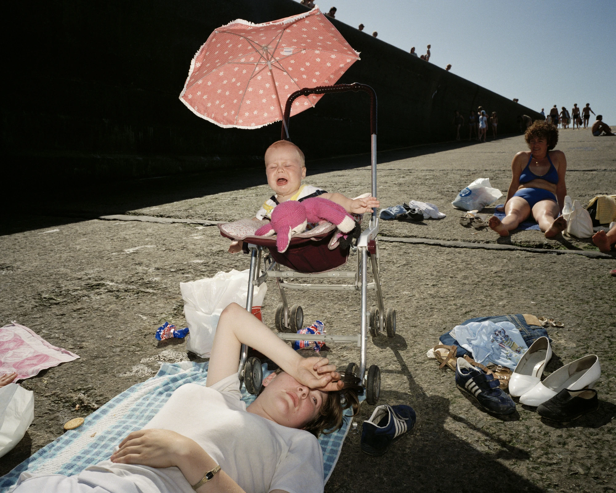 The Last Resort by Martin Parr, photographed by Martin Parr — A young mother lies on a blue checked towel with her forearm across her eyes while a baby cries in a pushchair beside her under a pink flowered parasol, shoes and clothes strewn over the…