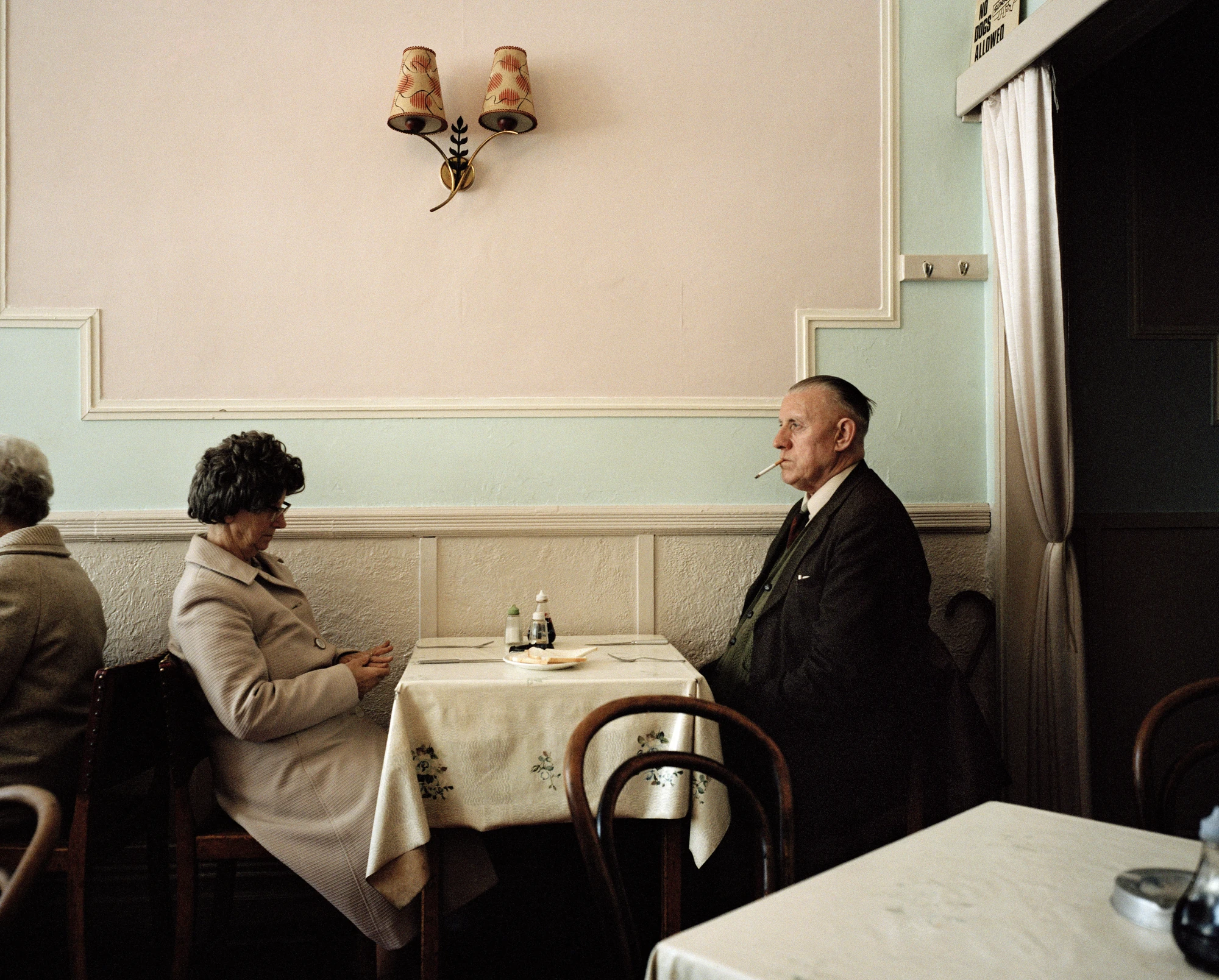 The Last Resort by Martin Parr, photographed by Martin Parr — In a cafe with pink and mint green walls, a woman in a cream coat sits with her hands folded and eyes down while a man in a dark suit smokes across the small table, a No Dogs Allowed sign by…