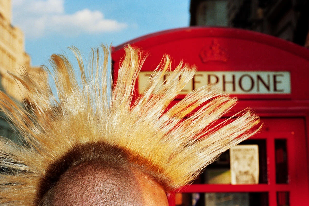 In Plain View by Martin Parr, photographed by Martin Parr — A bleached blond mohawk fills the foreground in front of a red London telephone box under a blue sky.