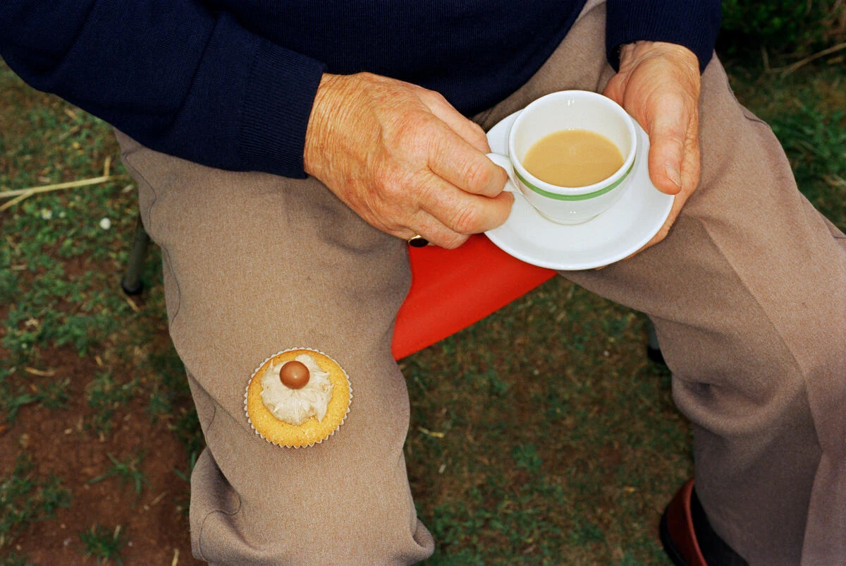 In Plain View by Martin Parr, photographed by Martin Parr — An elderly man in a navy jumper holds a cup of milky tea and its saucer over his knee, a cupcake in a paper case balanced on his thigh.