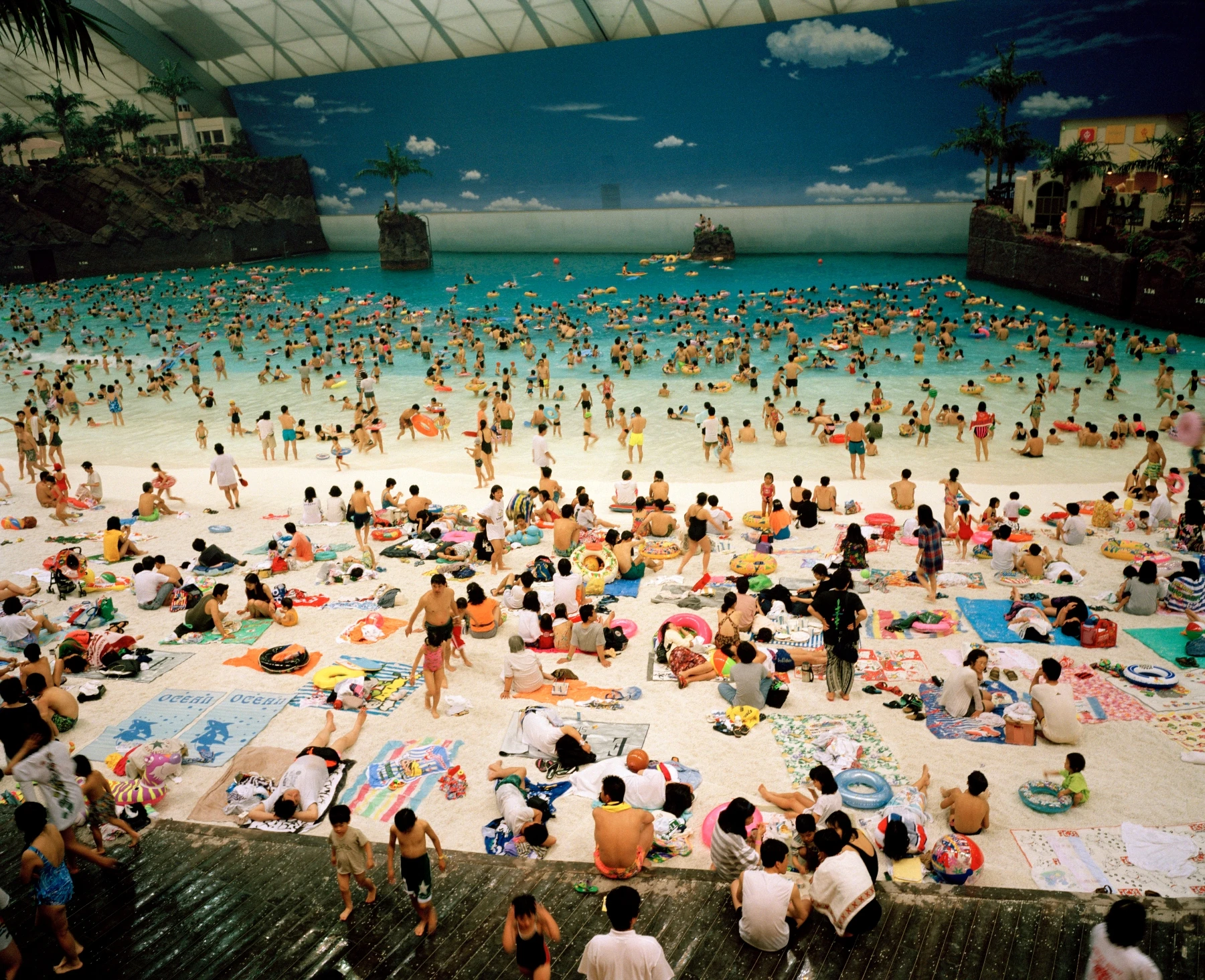 In Plain View by Martin Parr, photographed by Martin Parr — Crowds fill the white artificial sand and turquoise wave pool of an indoor beach resort beneath a painted sky.