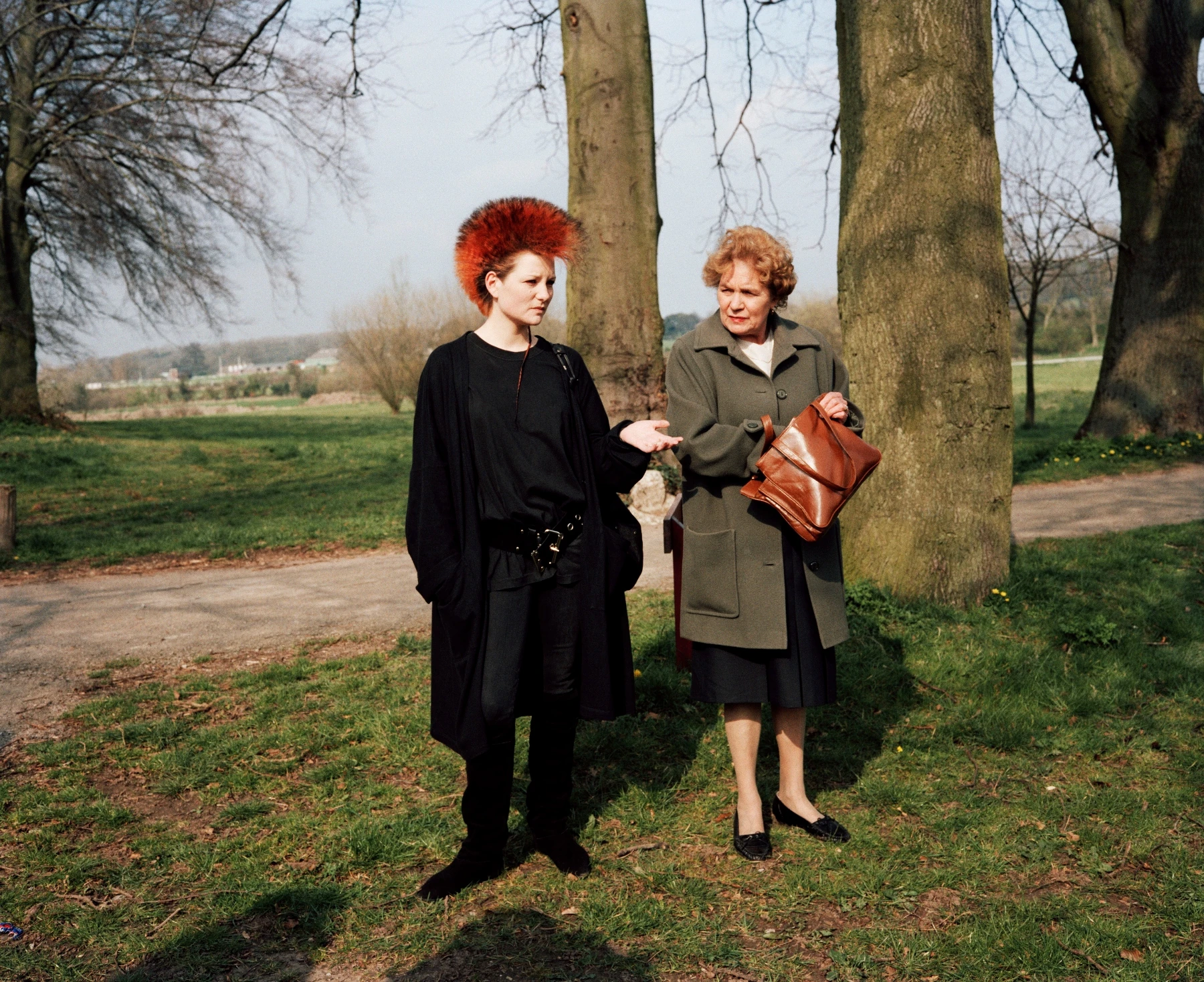 In Plain View by Martin Parr, photographed by Martin Parr — A young woman with an orange mohawk dressed in black stands beside an older woman in a green coat holding a tan handbag, bare trees behind.
