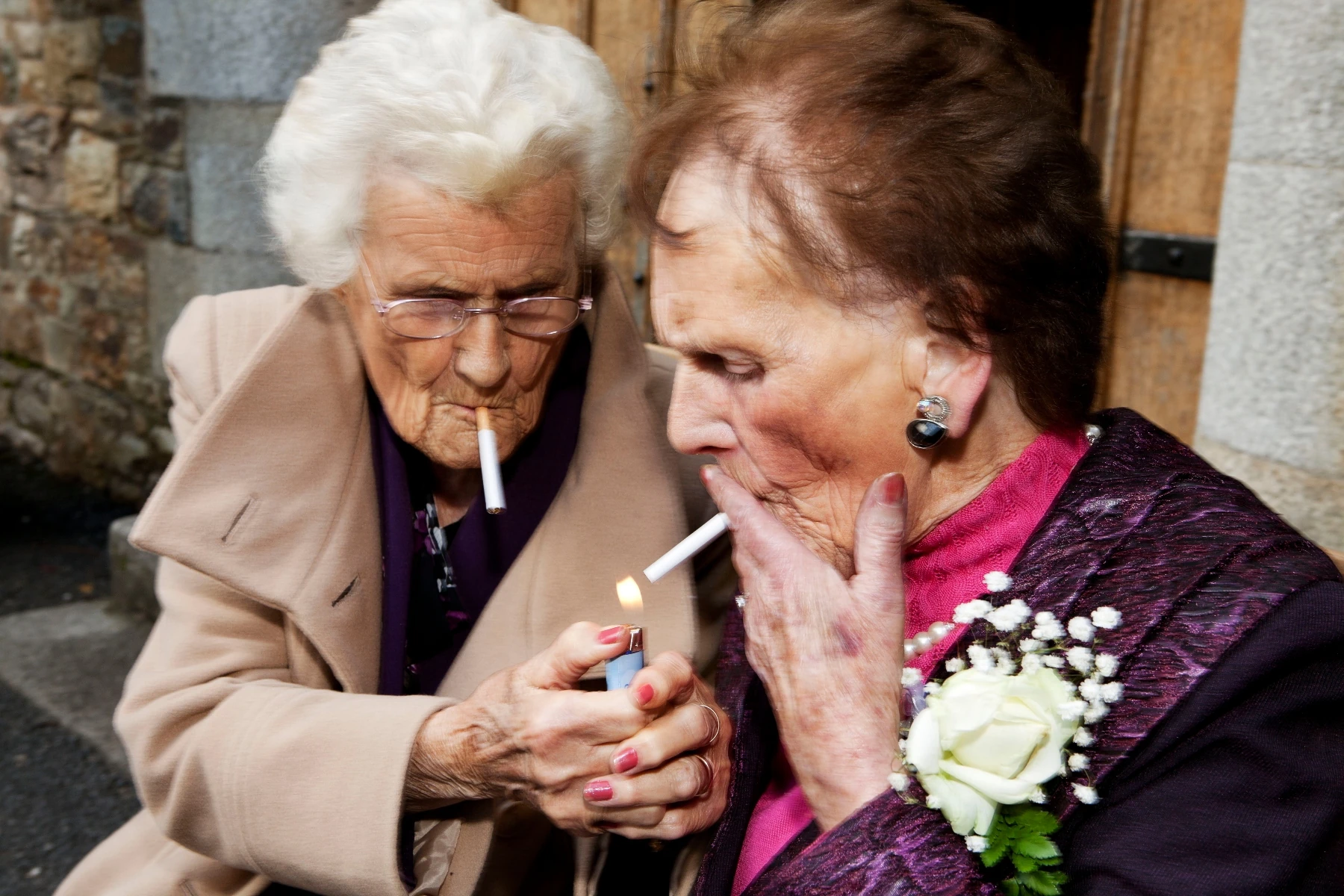 In Plain View by Martin Parr, photographed by Martin Parr — Two older women lean together outside a stone church, one lighting the other's cigarette, a white rose corsage pinned to a purple jacket.