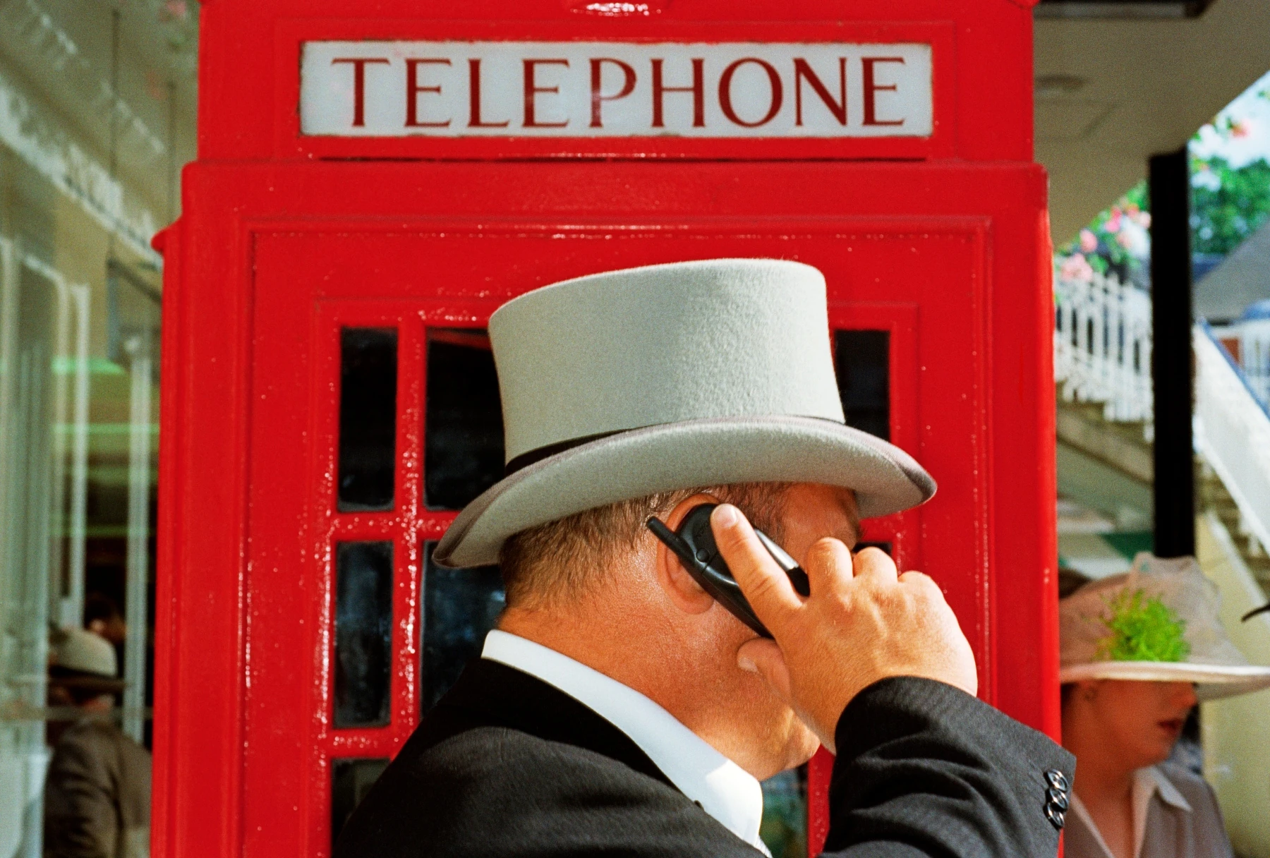 In Plain View by Martin Parr, photographed by Martin Parr — A man in a grey top hat presses a mobile phone to his ear in front of a red telephone box.