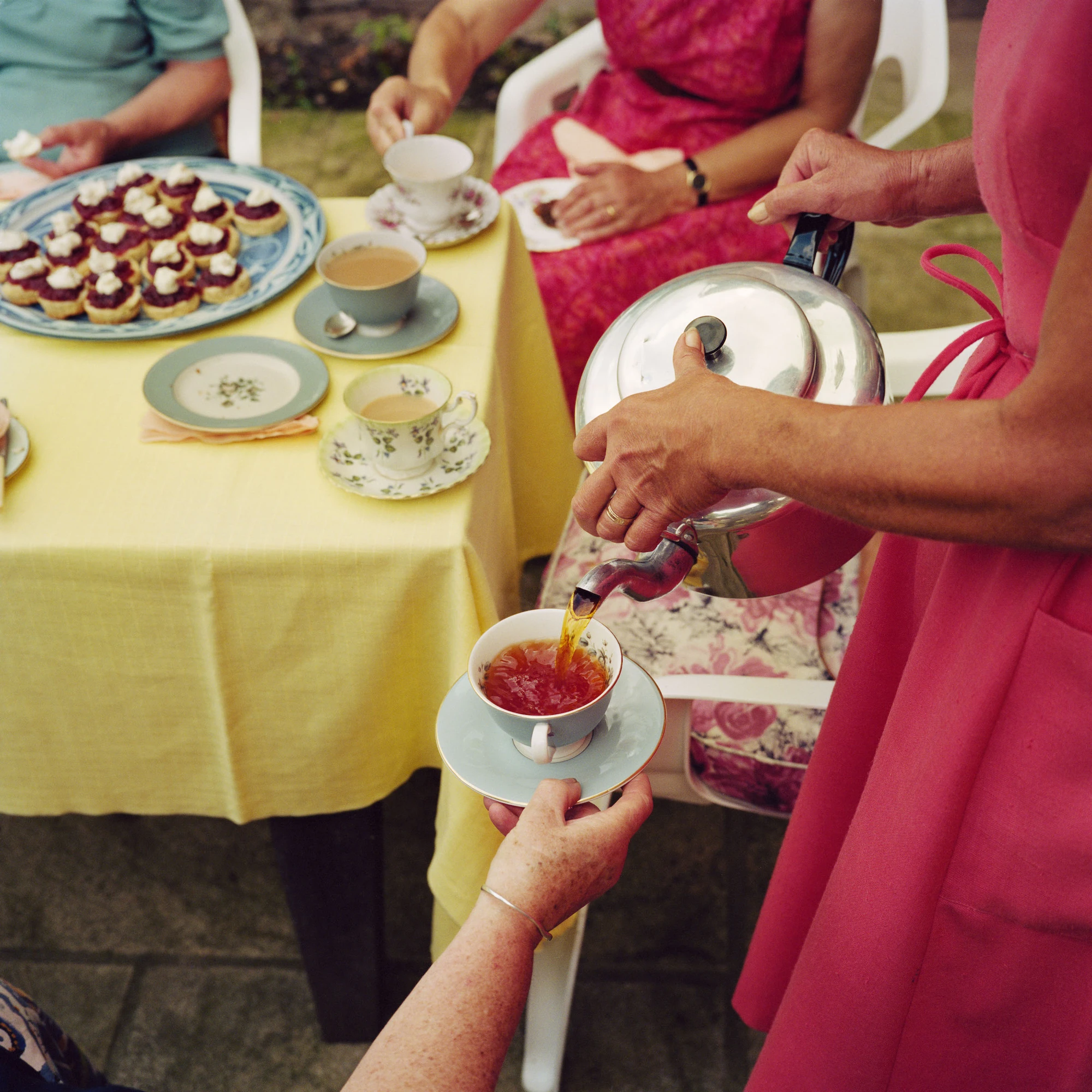 In Plain View by Martin Parr, photographed by Martin Parr — Tea pours from a chrome pot into a pale blue cup held over a saucer at a garden party, a yellow tablecloth set with jam tarts and floral china behind.