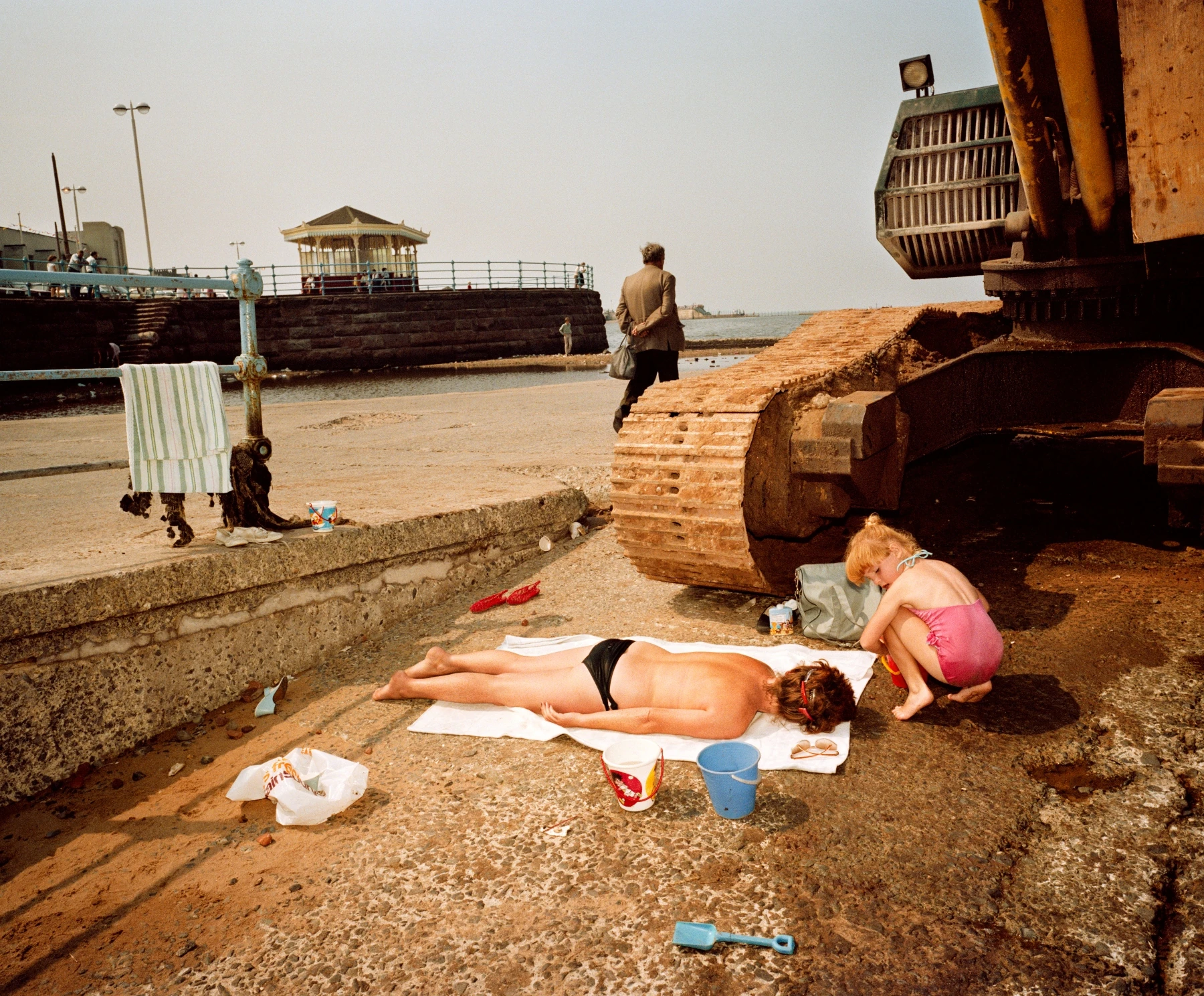 In Plain View by Martin Parr, photographed by Martin Parr — A woman sunbathes on a white towel on cracked concrete beside the rusted track of an excavator while a child crouches nearby.