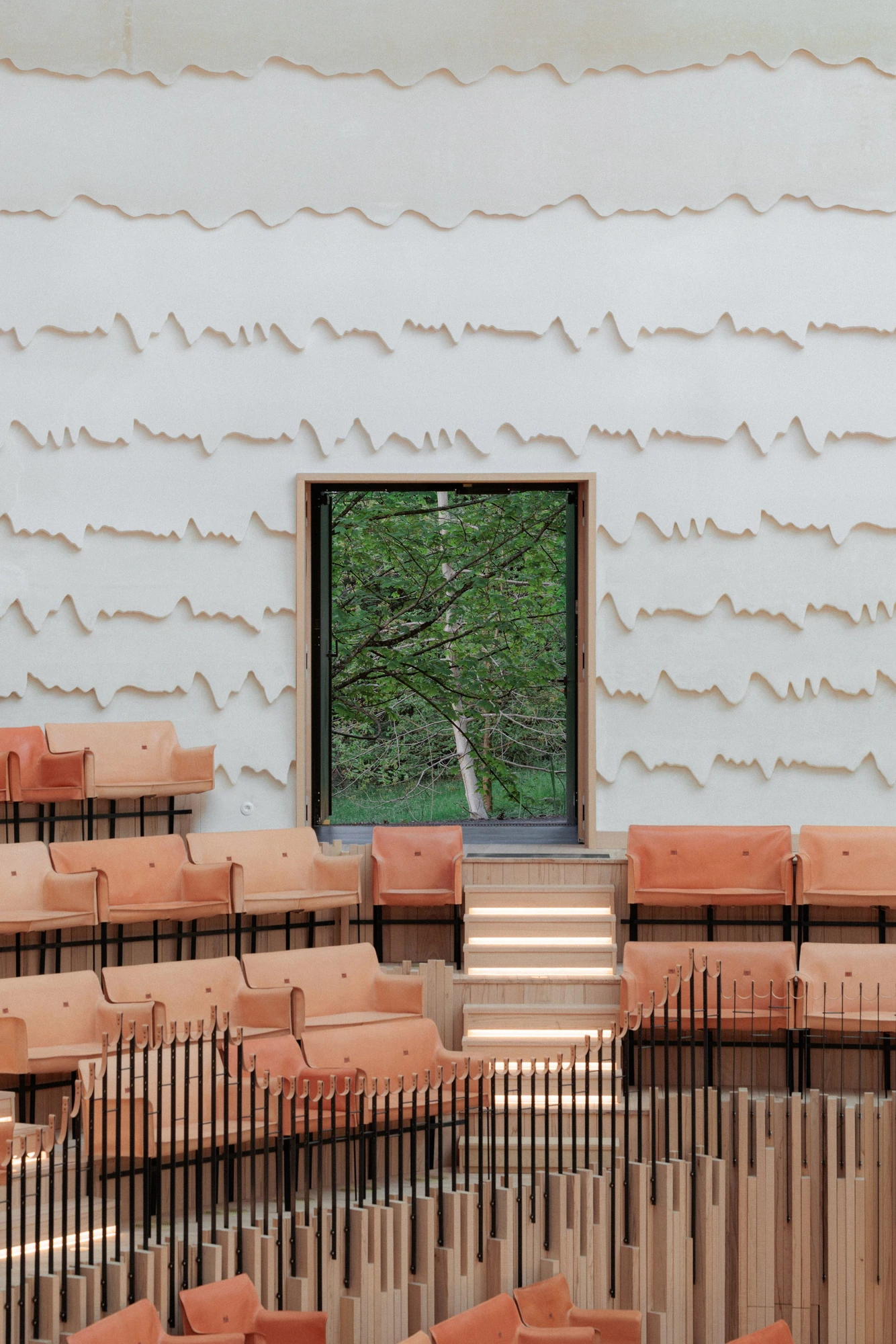 La Source Vive by PCA-Stream, photographed by PCA-Stream — Undulating plaster grooves running across the auditorium wall, broken by a tall window framing a birch in full leaf above the seating.