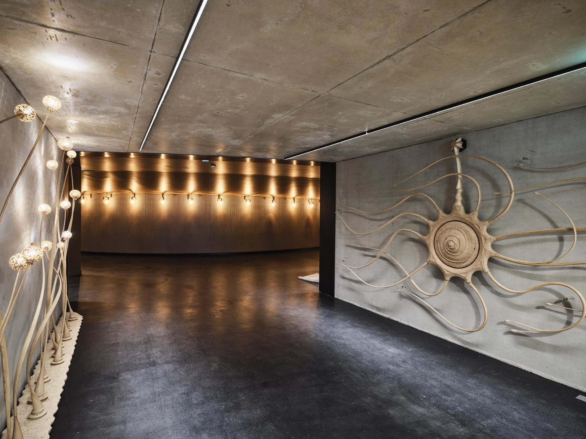 La Source Vive by PCA-Stream, photographed by PCA-Stream — Concrete-lined lobby with a woven rattan sun mounted on the wall and a lit, fluted timber bar counter in the room beyond.