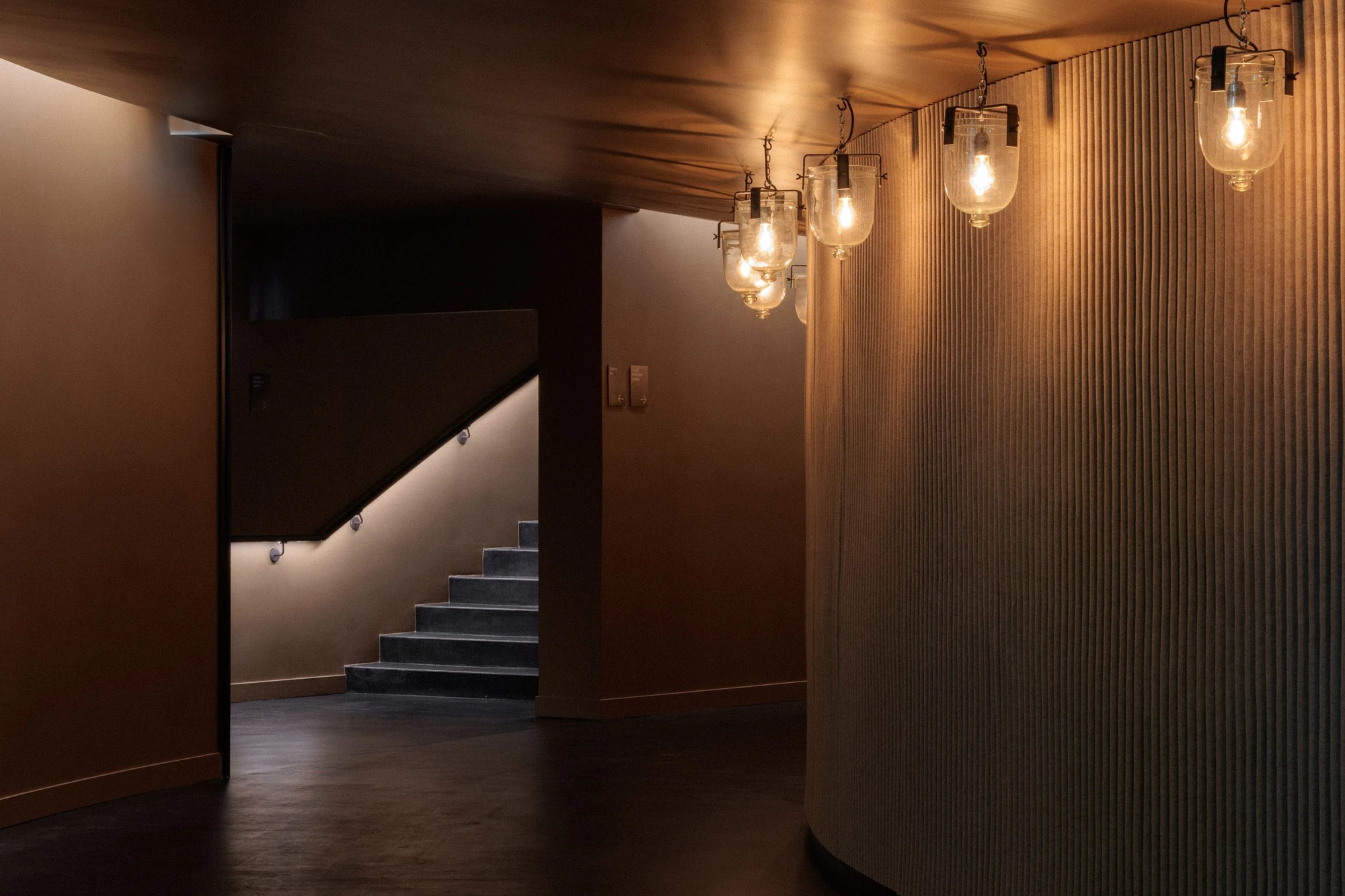 La Source Vive by PCA-Stream, photographed by PCA-Stream — Curved corridor lined in fluted plaster, hung with glass bell-jar pendants on chains, a lit stair visible beyond.