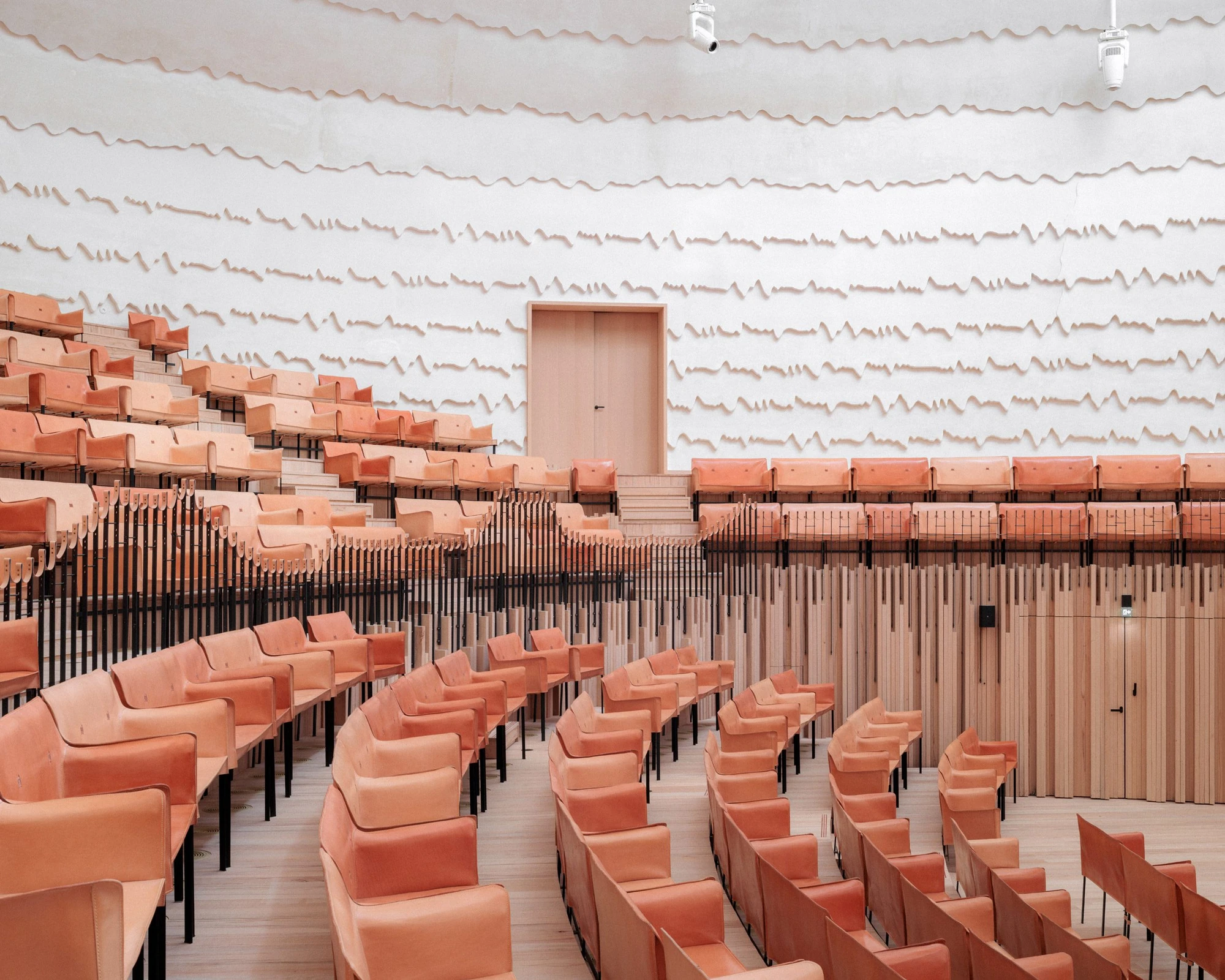 La Source Vive by PCA-Stream, photographed by PCA-Stream — Terraced rows of orange leather benches rising toward a timber door set into the grooved plaster wall.