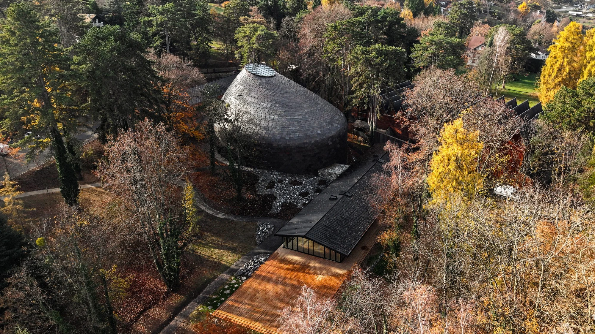 La Source Vive by PCA-Stream, photographed by PCA-Stream — Aerial view of the copper-tiled dome in an autumn woodland clearing, with a glazed pavilion and timber terrace running downhill from it.