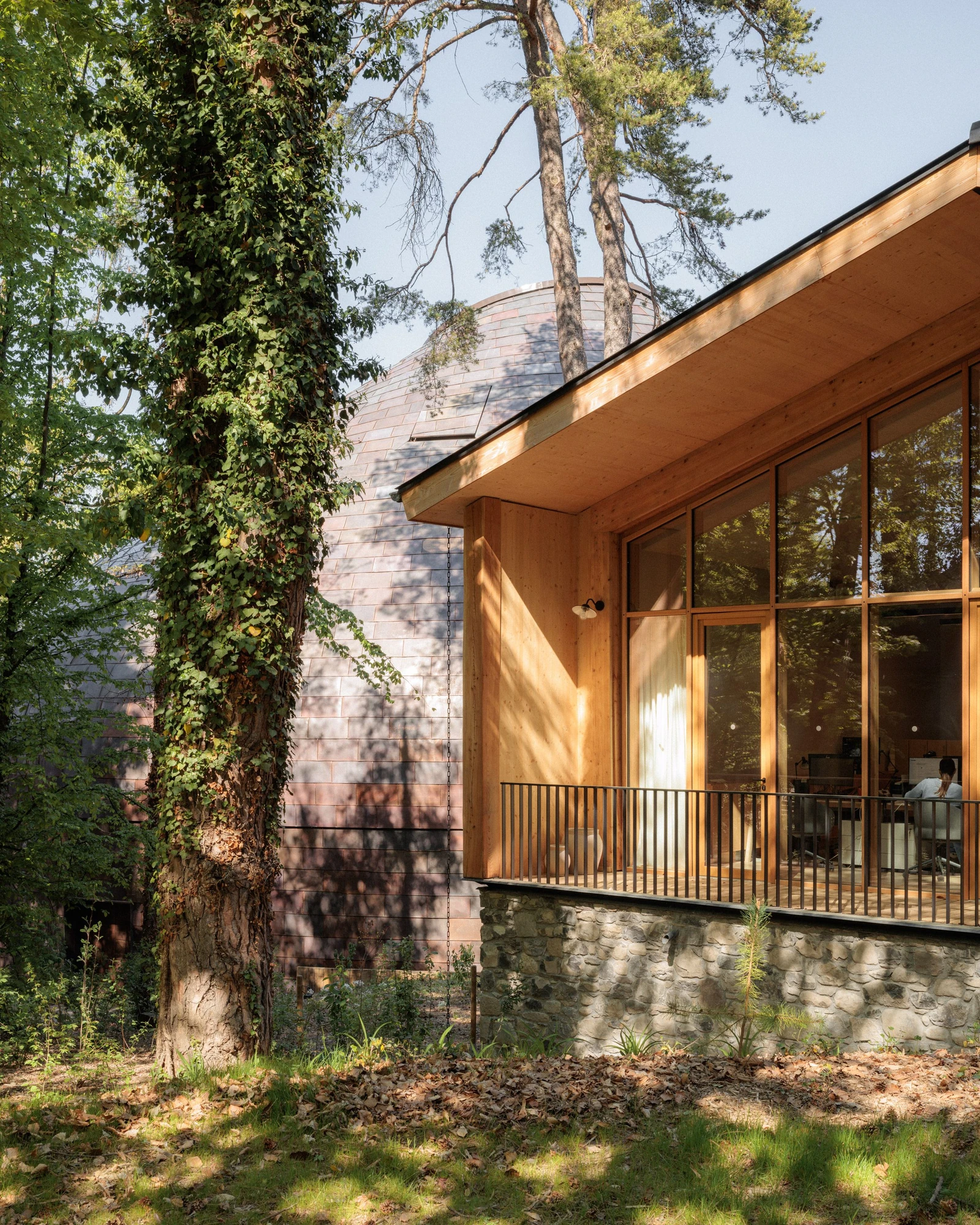 La Source Vive by PCA-Stream, photographed by PCA-Stream — The larch-clad backstage building on a rubble stone base, the copper dome rising behind and a figure at a desk inside.