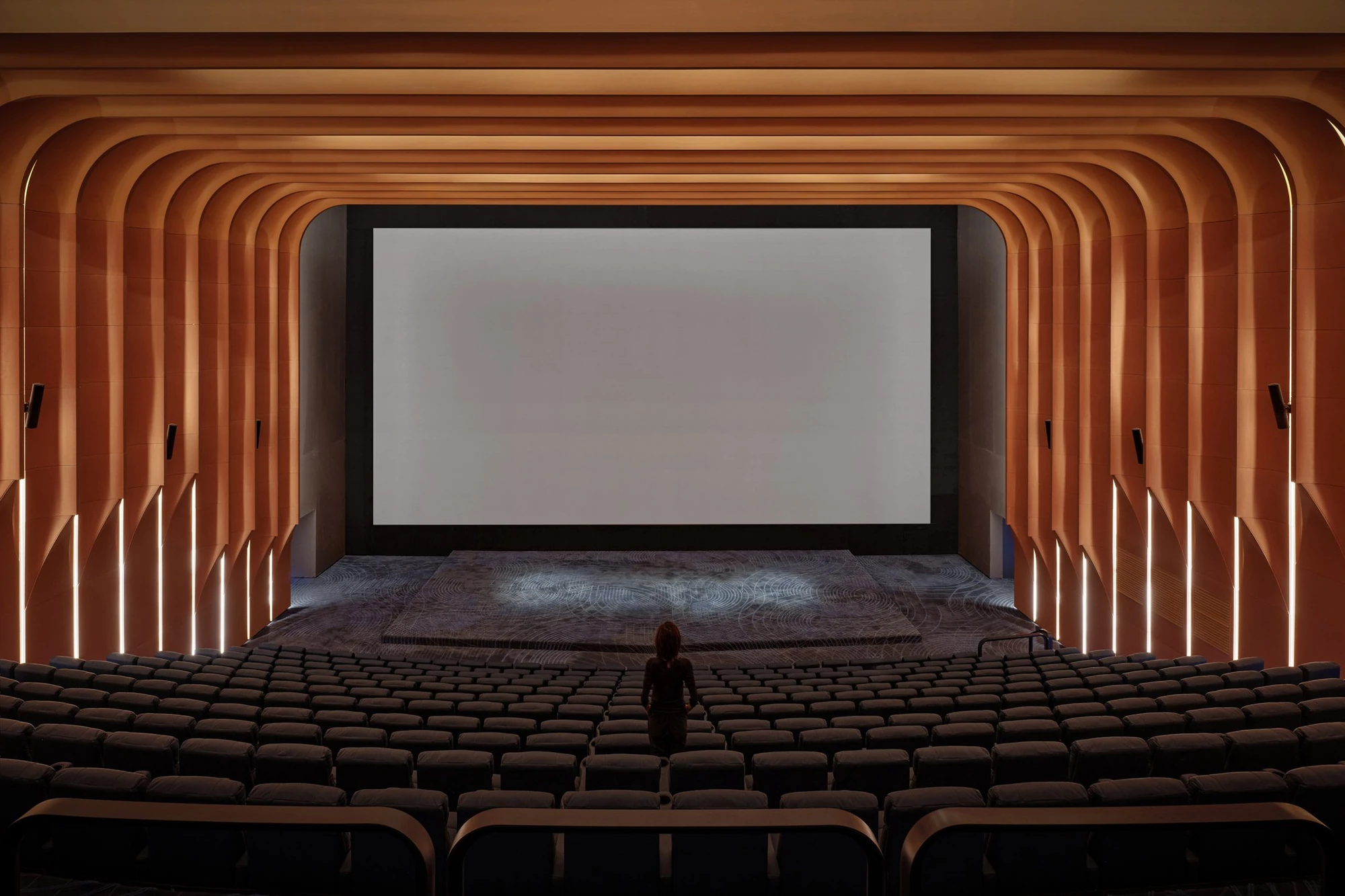 Haikou Gaoxingli Insun Cinema by One Plus Partnership, photographed by One Plus Partnership — The main auditorium, the screen framed by curving wooden bands with light set between them and a single figure standing among the seats.