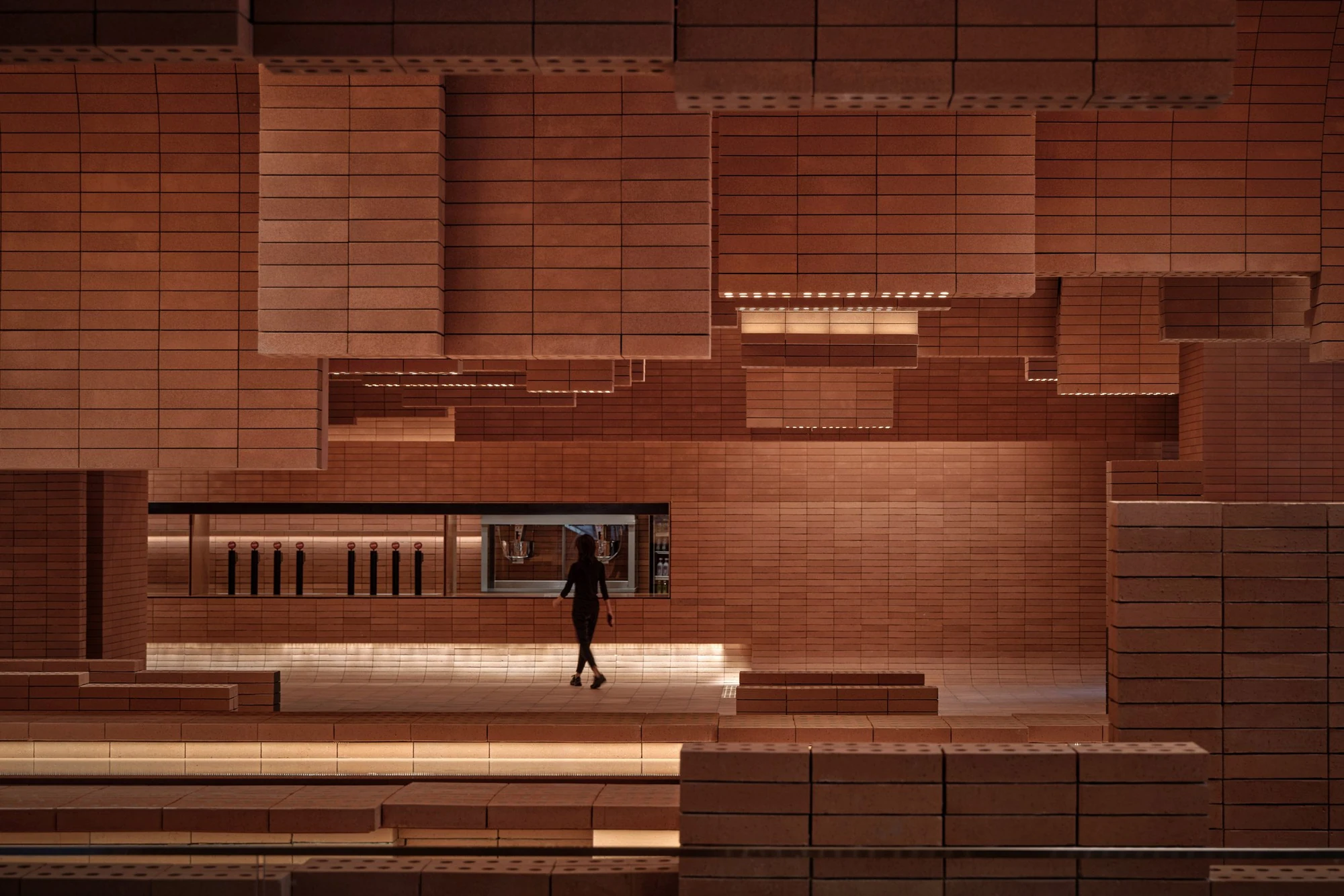 Haikou Gaoxingli Insun Cinema by One Plus Partnership, photographed by One Plus Partnership — Frontal view of the concession window, a figure crossing the brick floor beneath stepped brick blocks that hang from the ceiling with light strips along their…