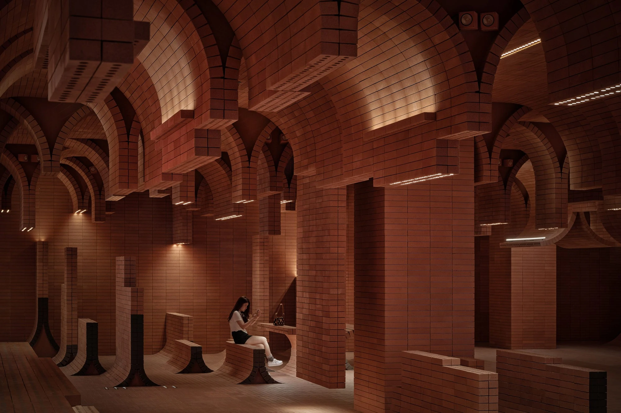 Haikou Gaoxingli Insun Cinema by One Plus Partnership, photographed by One Plus Partnership — A woman sits with her phone on a curved brick bench under lit brick arches, with linear light strips set into the overhanging slabs.