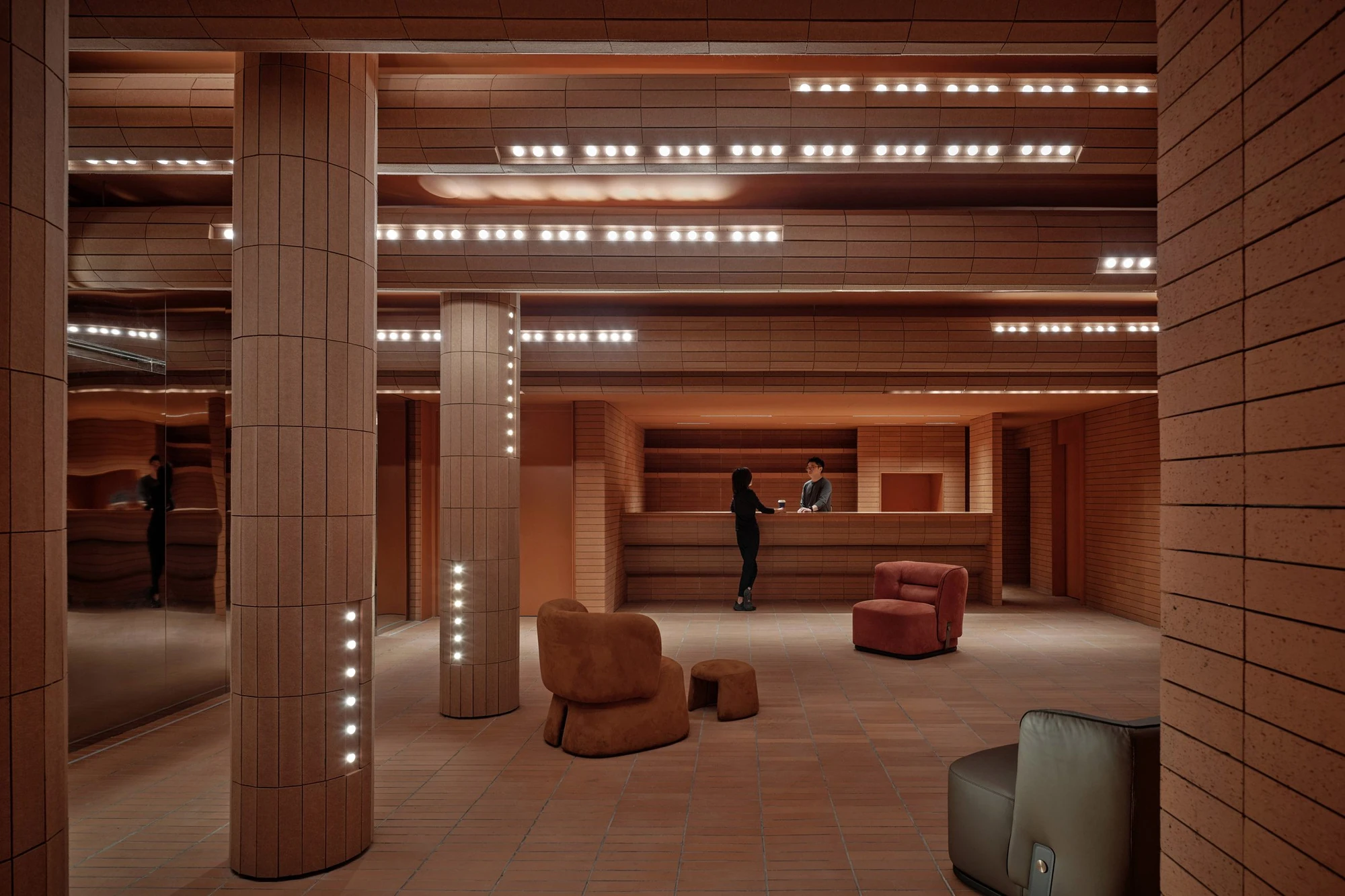 Haikou Gaoxingli Insun Cinema by One Plus Partnership, photographed by One Plus Partnership — Tiled terracotta columns studded with bulbs stand in a lounge, upholstered chairs on the tiled floor and two people at a counter beyond.