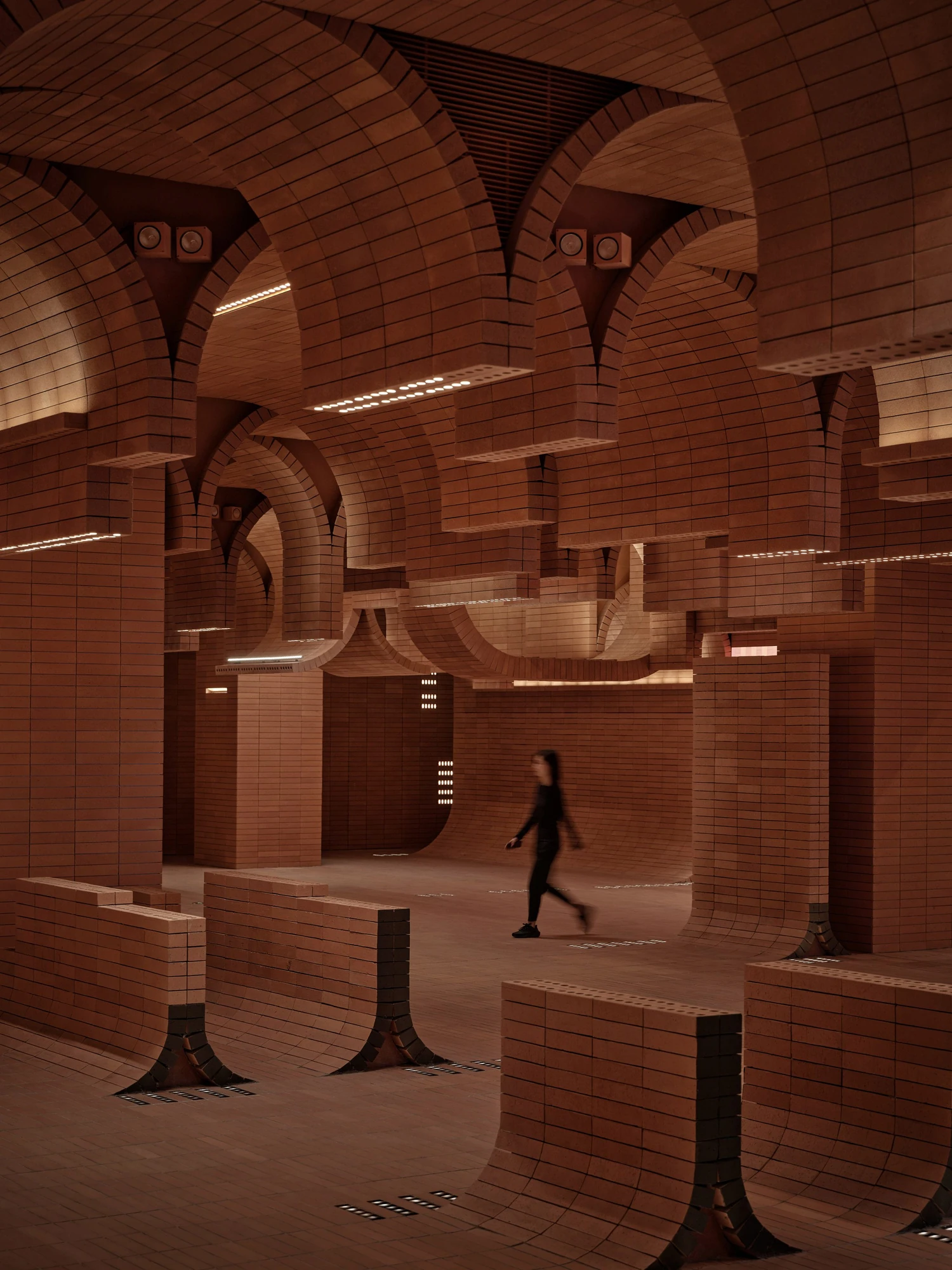 Haikou Gaoxingli Insun Cinema by One Plus Partnership, photographed by One Plus Partnership — A figure walks through the brick arcade, speakers mounted in the vaults and small square floor lights marking the route between curved brick benches.