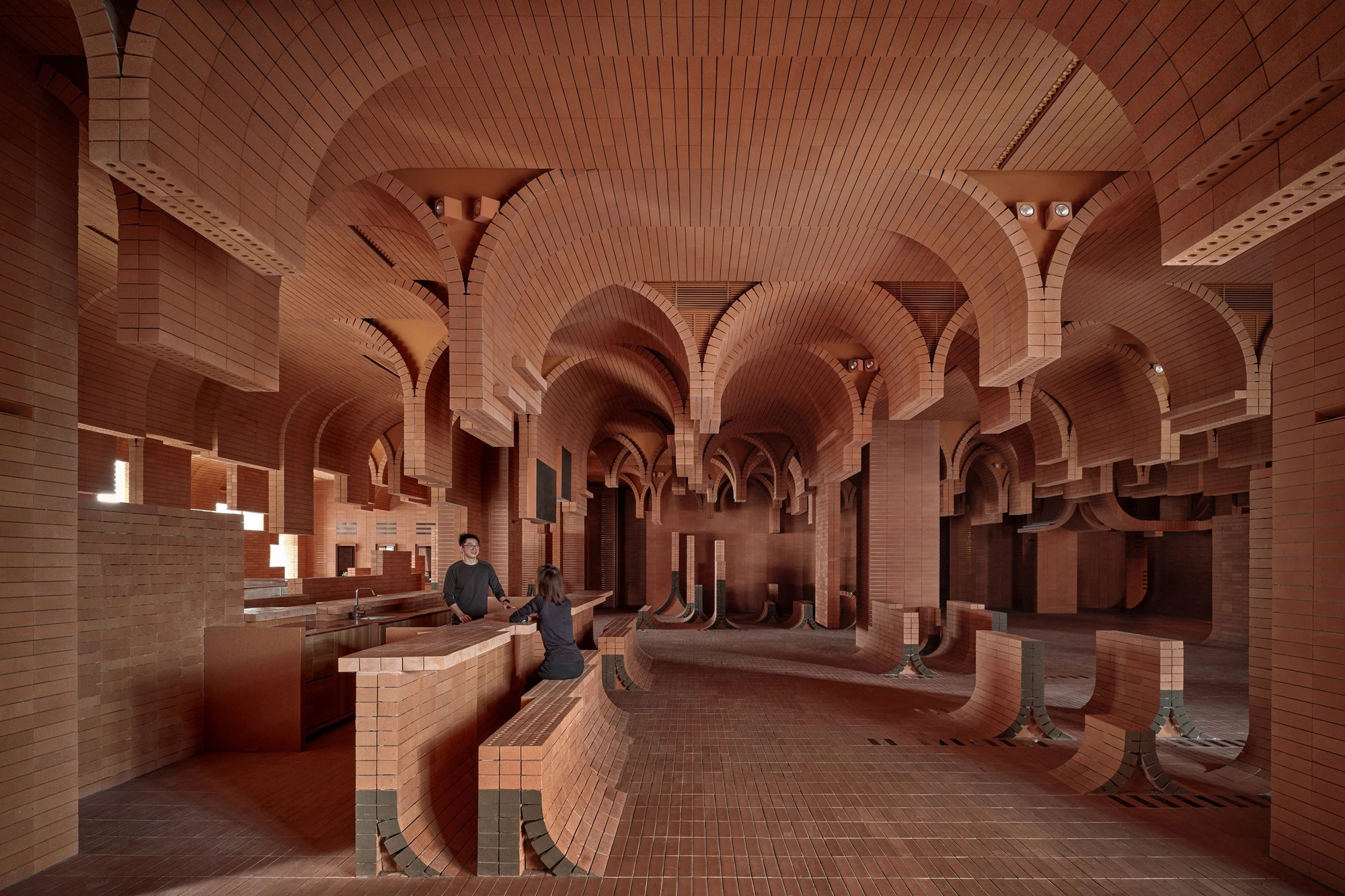 Haikou Gaoxingli Insun Cinema by One Plus Partnership, photographed by One Plus Partnership — Two people at the cafe counter under a run of brick vaults, curved brick benches lining the floor with grey painted ends.