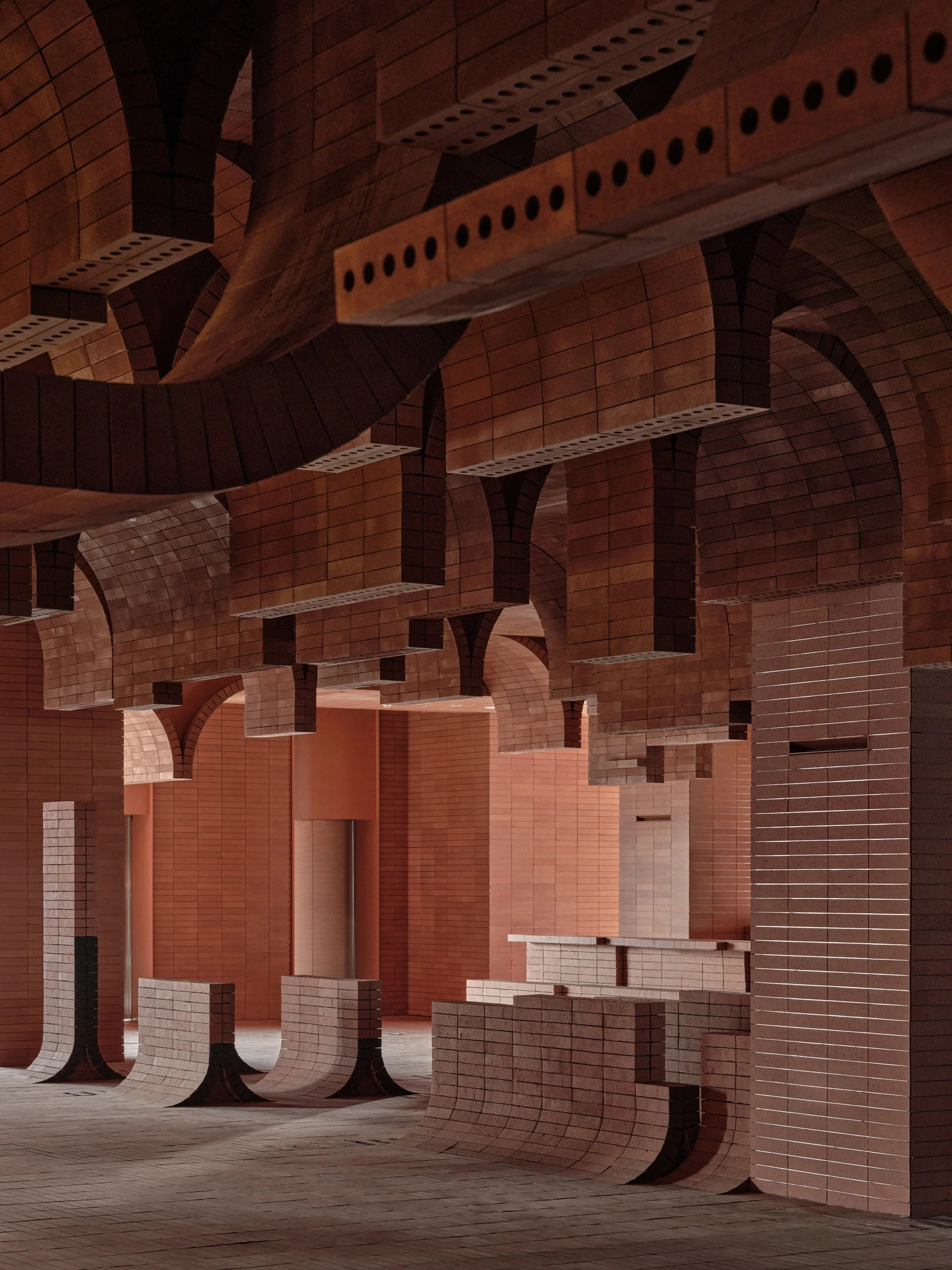 Haikou Gaoxingli Insun Cinema by One Plus Partnership, photographed by One Plus Partnership — Suspended brick beams with rows of round perforations run above curved brick counters, the far walls washed a pale salmon by concealed light.
