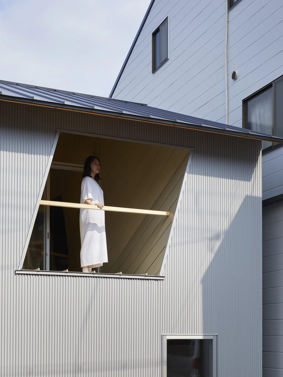 24mm Plywood House by Alphaville Architects, photographed by Alphaville Architects — Looking up at the corrugated grey facade, where a triangular window hinges outward and a woman in white stands behind a slim timber mullion against plywood boarding,…