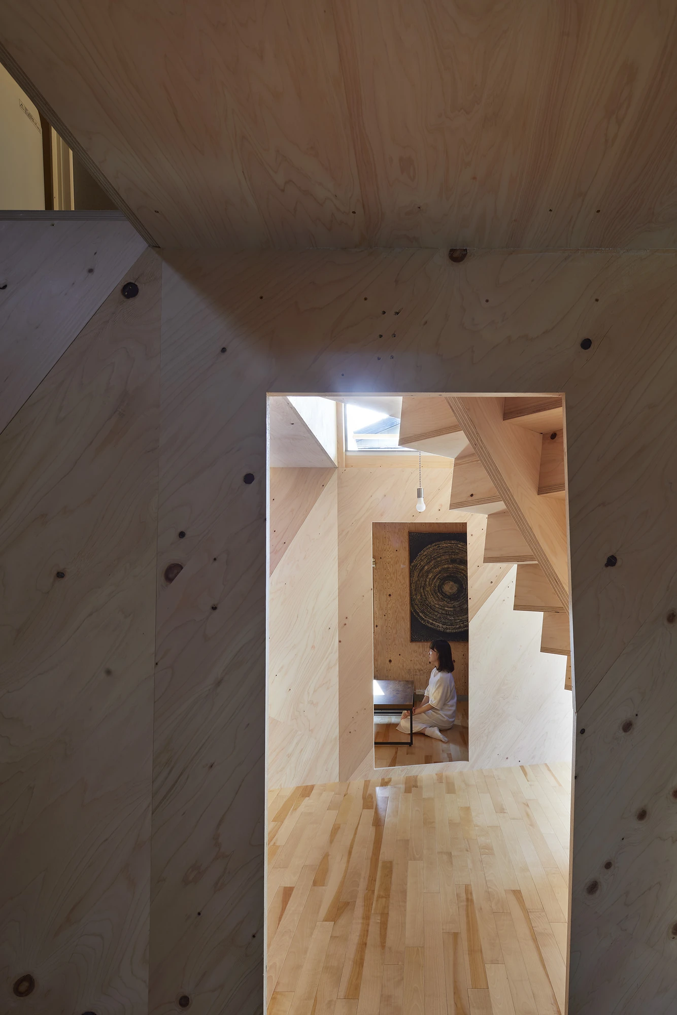 24mm Plywood House by Alphaville Architects, photographed by Alphaville Architects — Looking up through the atrium at plywood soffits and a slim timber post, past a glazed opening onto the terrace where a figure in white passes, with a square glass…