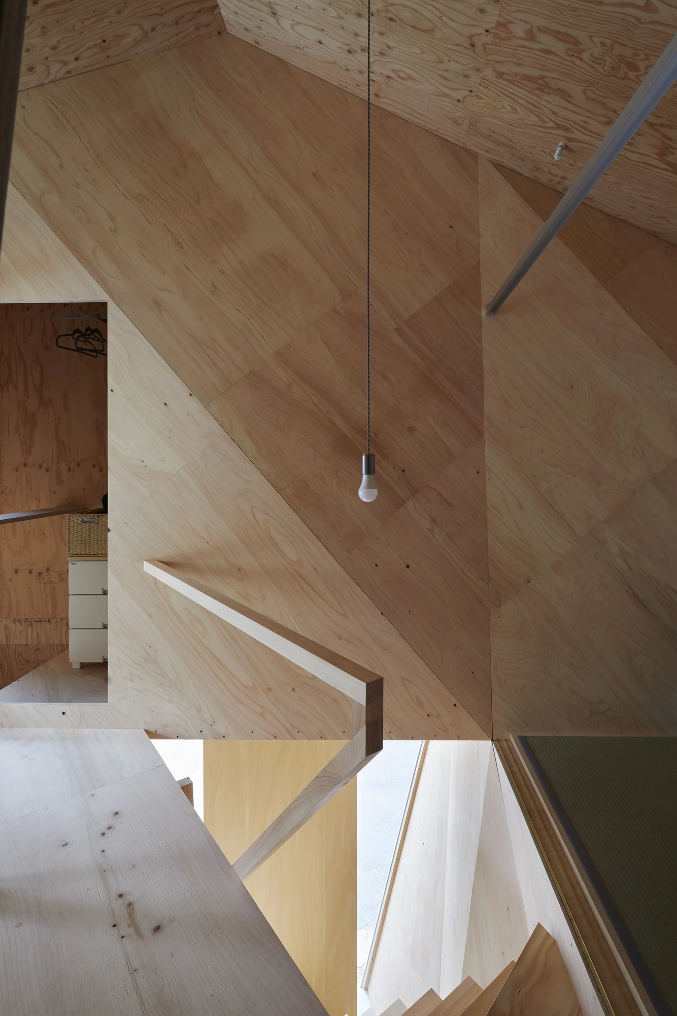 24mm Plywood House by Alphaville Architects, photographed by Alphaville Architects — Angled plywood ceiling and wall planes converging above a tatami-edged opening, a single bare bulb on a braided cord and a slim timber handrail cantilevered from the…