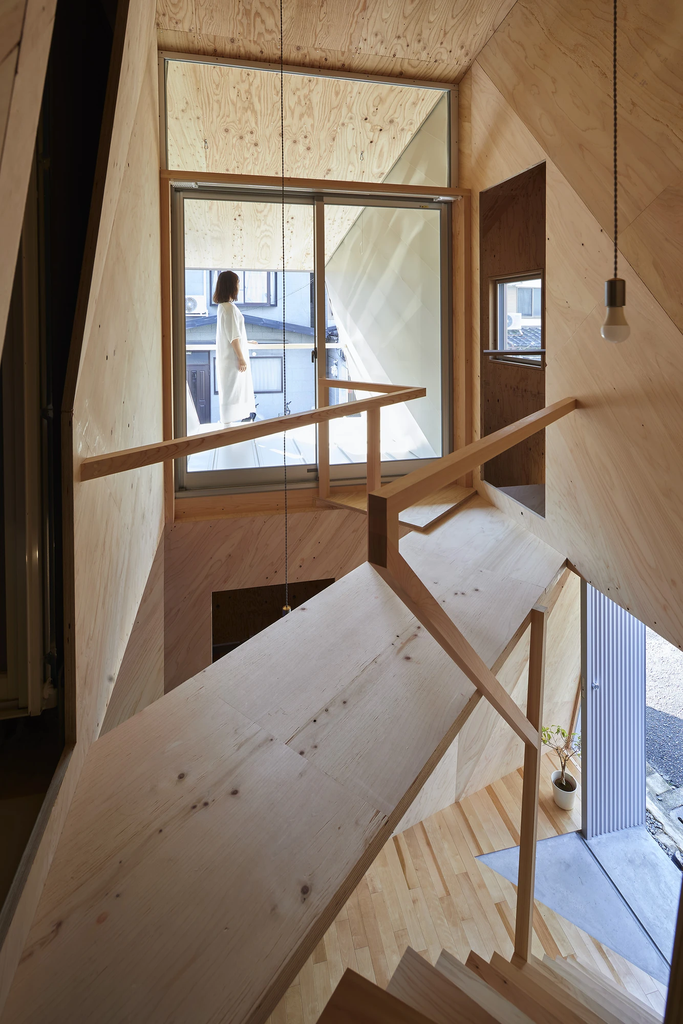 24mm Plywood House by Alphaville Architects, photographed by Alphaville Architects — Looking straight down through a rectangular floor opening onto the plywood stair and a figure seated on the ground floor beside a large dark circular painting, sunlight…