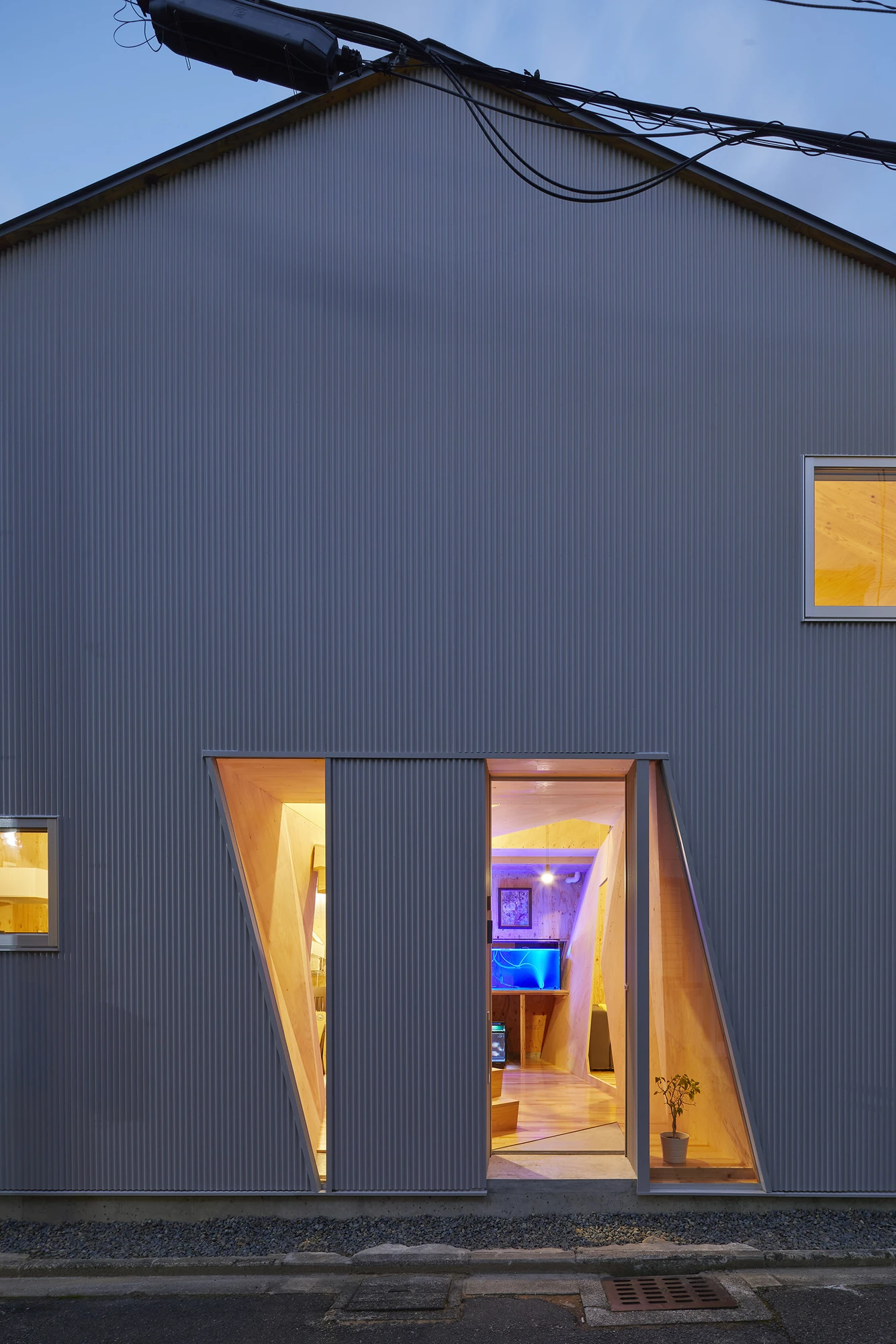 24mm Plywood House by Alphaville Architects, photographed by Alphaville Architects — The grey corrugated wall at dusk, with two angular openings glowing amber and blue and revealing plywood-lined rooms, a concrete boundary wall at the left edge and…