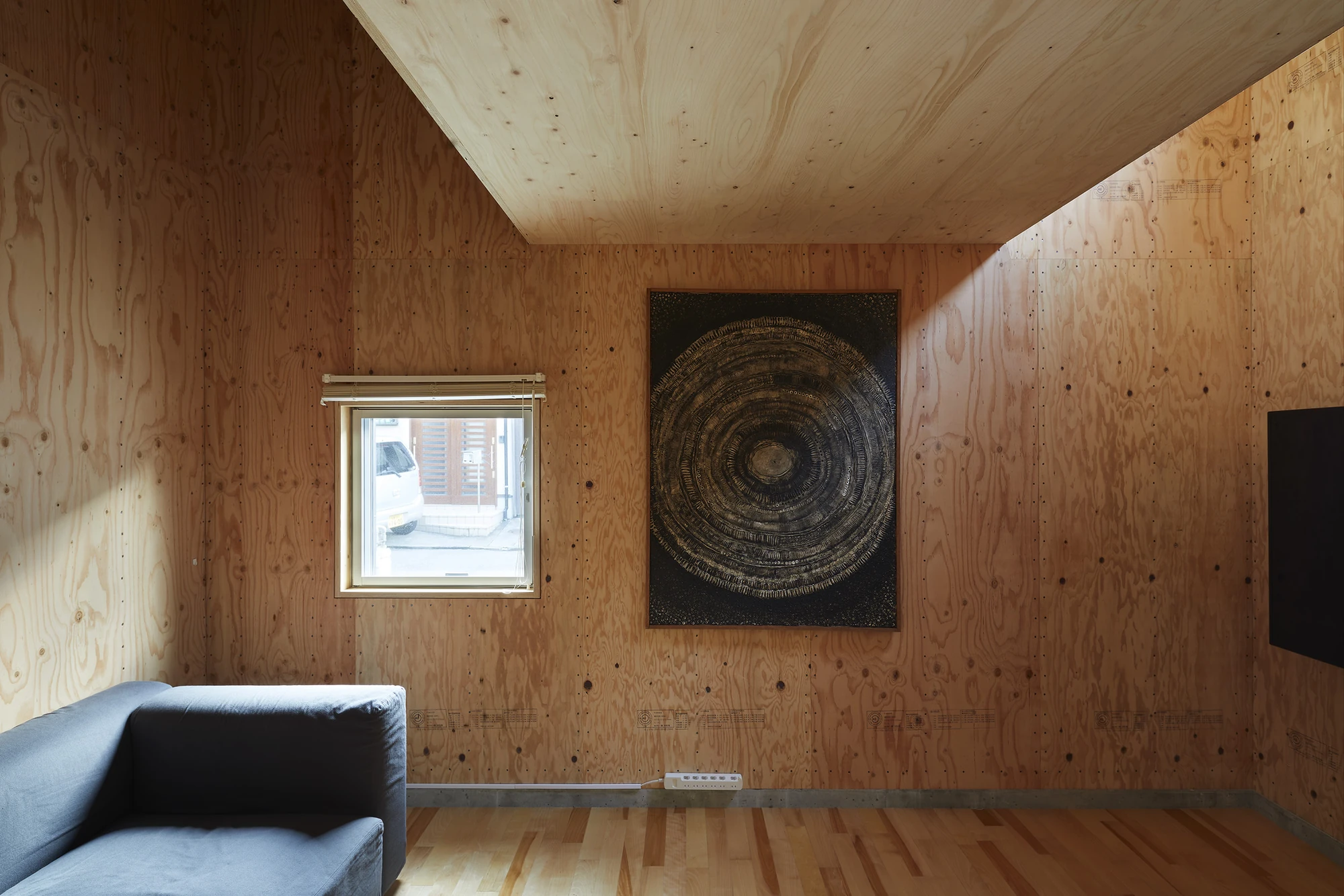 24mm Plywood House by Alphaville Architects, photographed by Alphaville Architects — Living space with reddish plywood walls carrying visible mill stamps and screw lines, a dark circular painting hung between a small square window and a grey sofa on a…