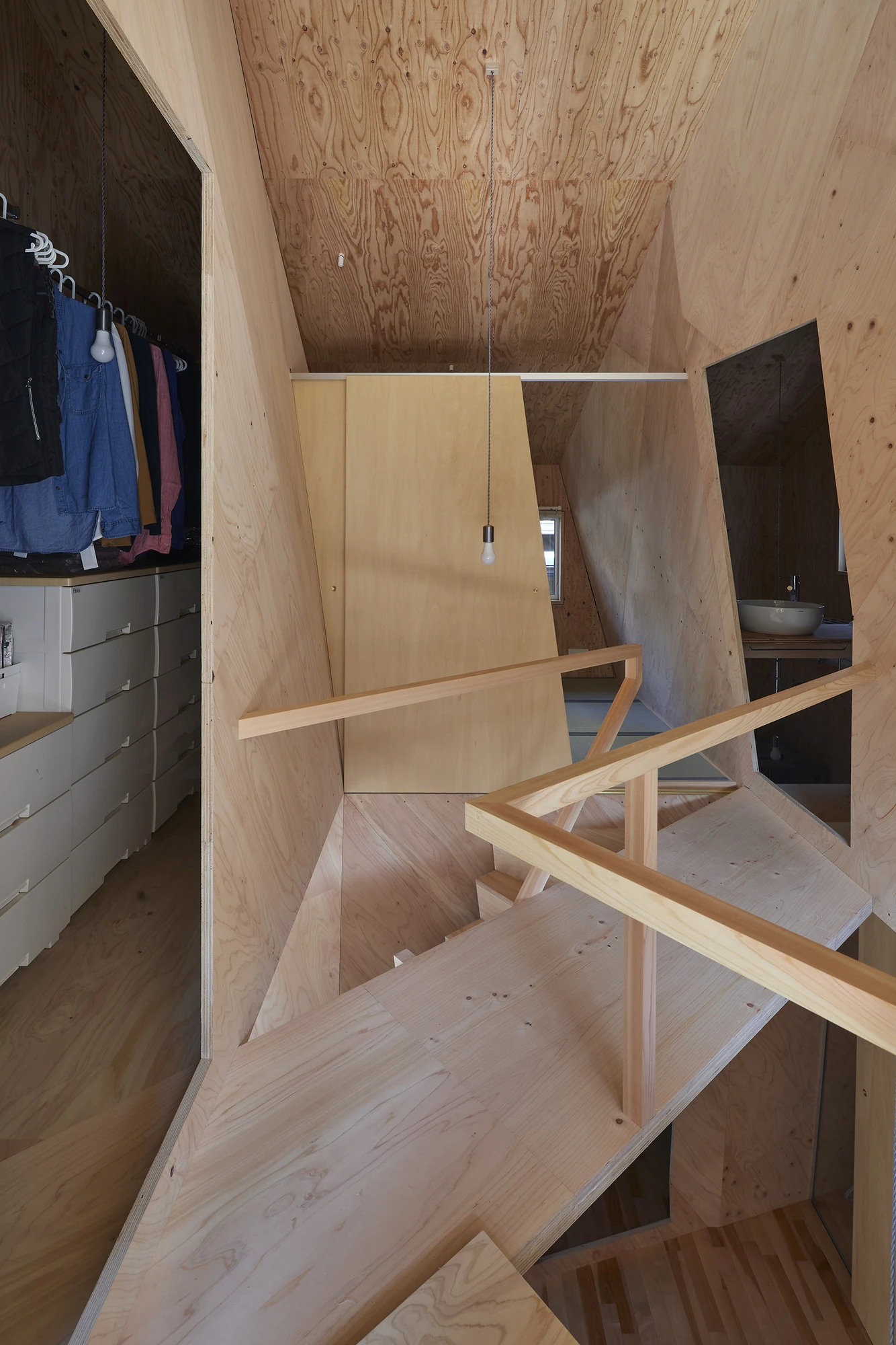 24mm Plywood House by Alphaville Architects, photographed by Alphaville Architects — Upper-level view across folded plywood planes and slender timber bracing towards an open storage recess where clothes hang on a rail, a bare bulb strung on a cable…