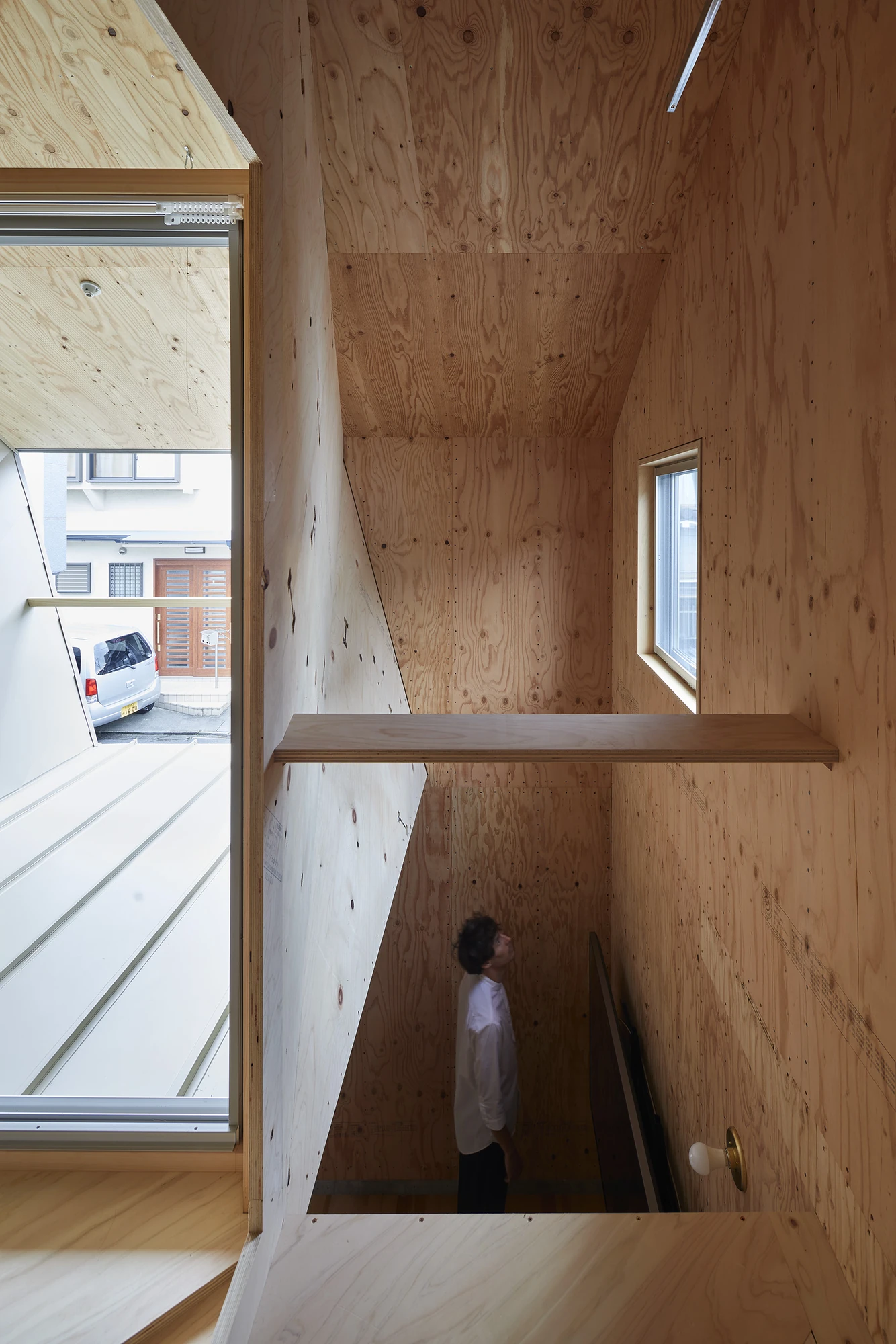 24mm Plywood House by Alphaville Architects, photographed by Alphaville Architects — Upper room lined entirely in structural plywood, with a glazed hatch set flush into the floor, a full-width window looking onto the street and a figure moving behind a…