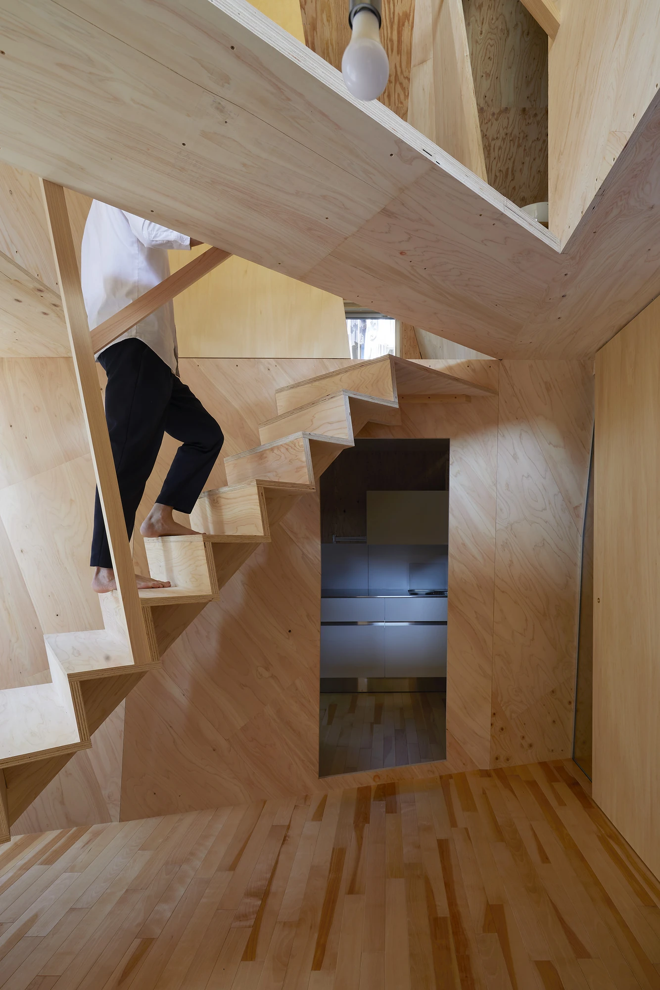 24mm Plywood House by Alphaville Architects, photographed by Alphaville Architects — A person in black trousers and white shirt descends the open plywood stair barefoot, passing a rectangular cut in the folded partition that frames a stainless kitchen…