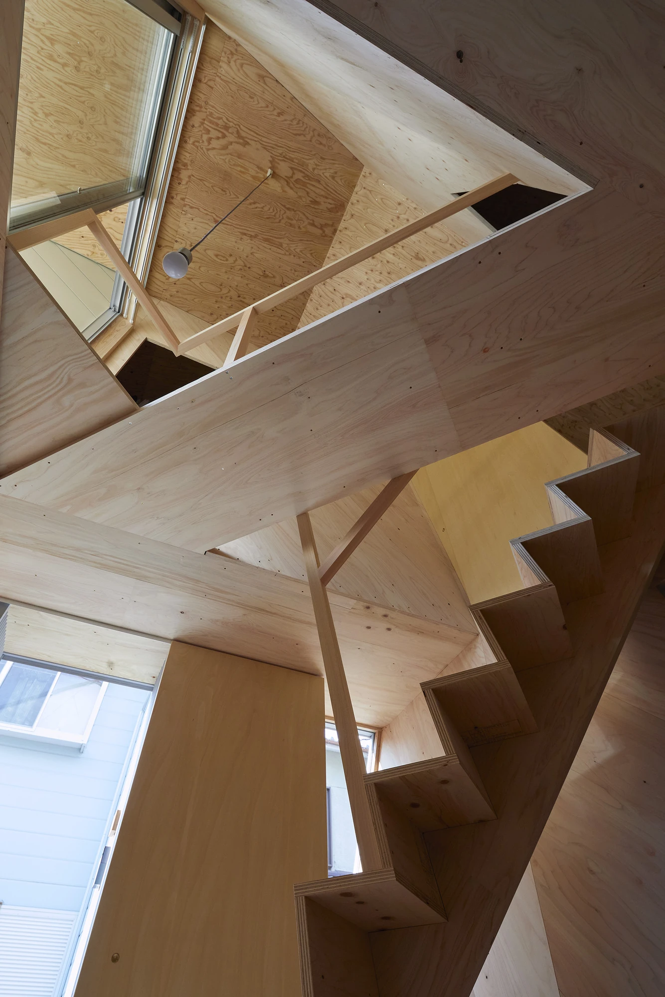 24mm Plywood House by Alphaville Architects, photographed by Alphaville Architects — Upward view into the folded plywood partition, its angled planes and screw lines meeting overhead while the stair soffit cuts across and a bare bulb hangs on a twisted…