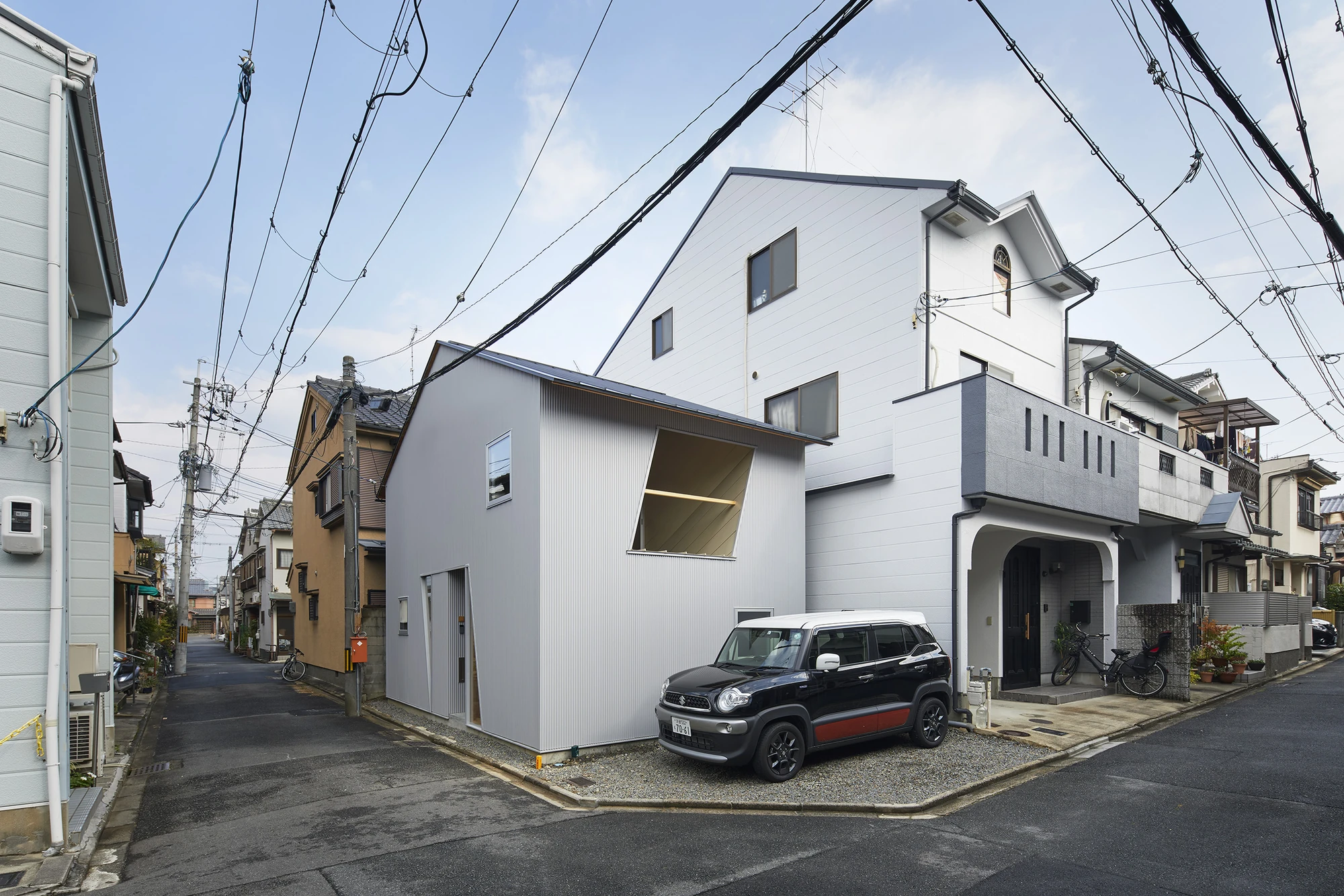 24mm Plywood House by Alphaville Architects, photographed by Alphaville Architects — The house seen from a road junction between neighbouring houses, its grey corrugated volume set on gravel with a car alongside, a hinged triangular flap open on the…