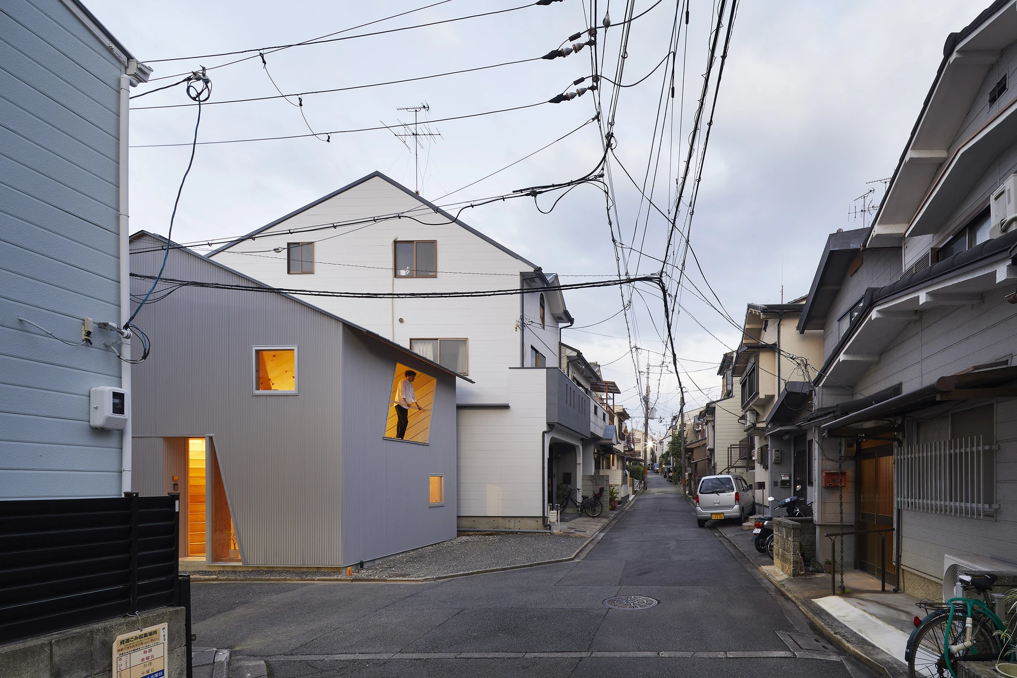 24mm Plywood House by Alphaville Architects, photographed by Alphaville Architects — The house at dusk from the street, its lit trapezoidal entrance and upper window casting warm plywood light into the lane, a figure standing in the opening beneath a…