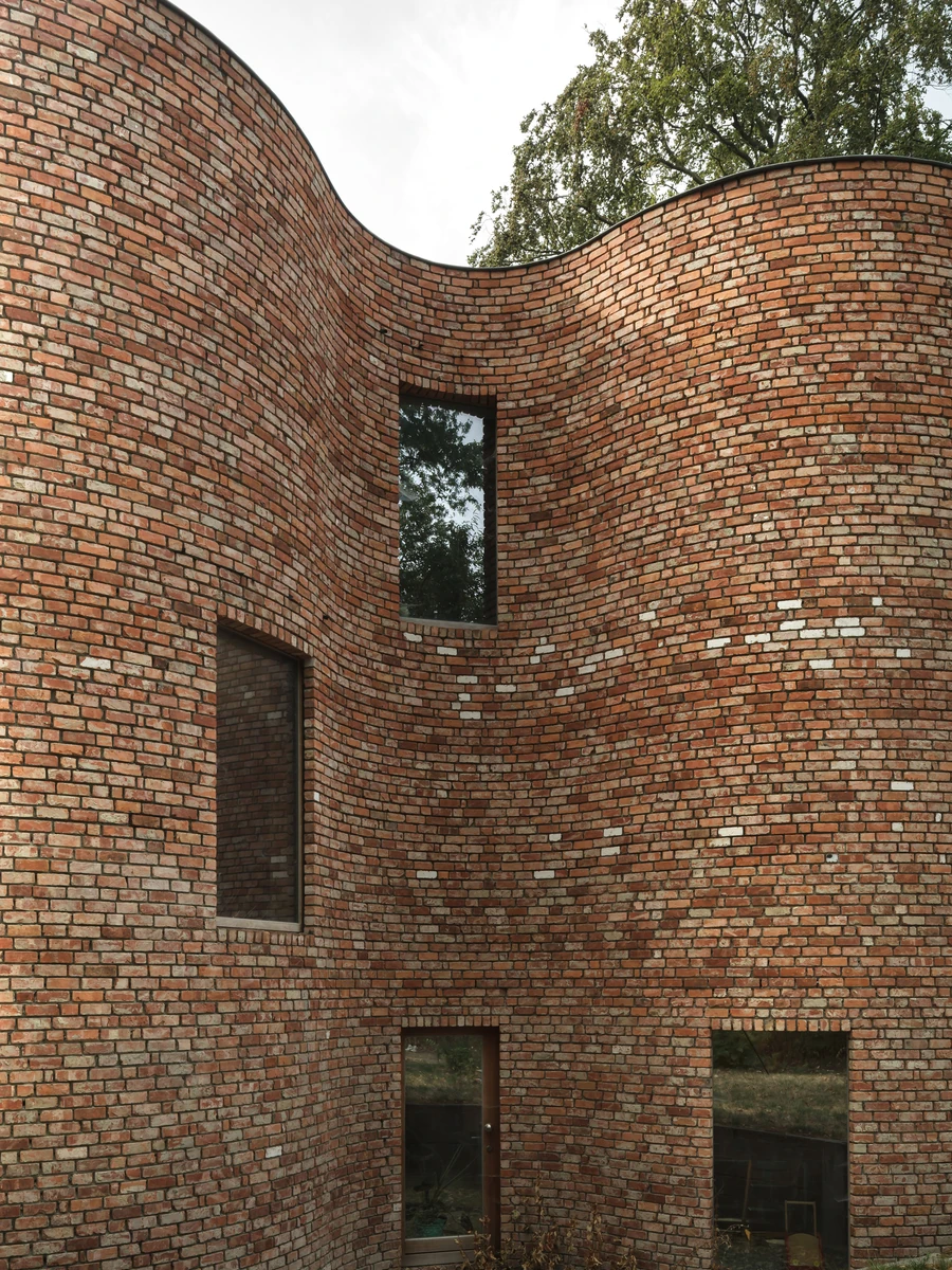 GjG House by BLAF Architecten, photographed by BLAF Architecten — The reclaimed brick wall curving in and out, its parapet drawn as an S against white sky, with a narrow slot window and a glazed door at the base.