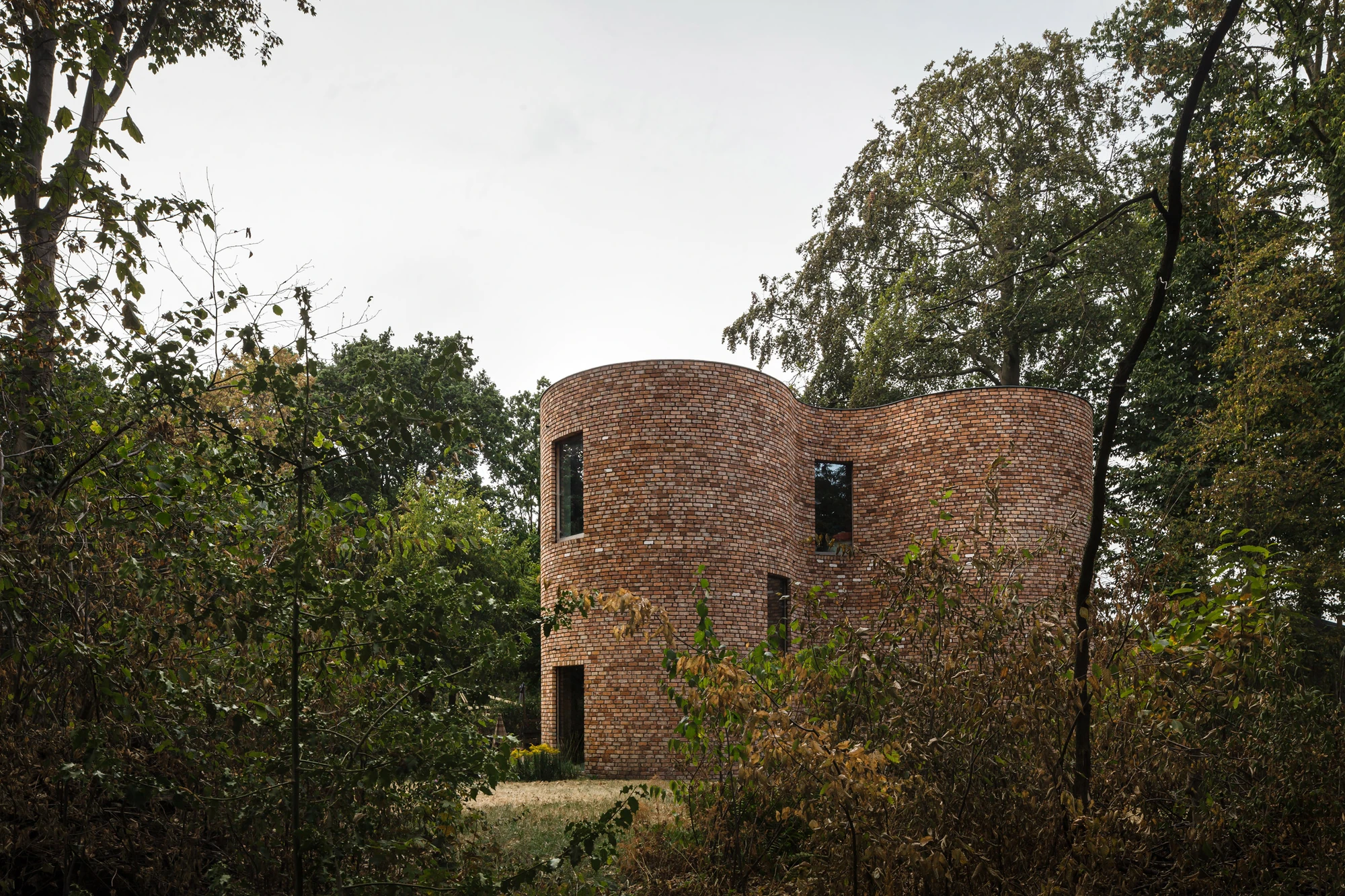 GjG House by BLAF Architecten, photographed by BLAF Architecten — The full silhouette of the house under overcast sky, the curved brick body framed by trees and scrub on both sides.