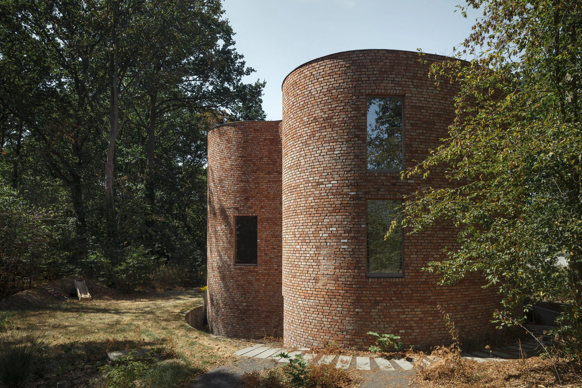 GjG House by BLAF Architecten, photographed by BLAF Architecten — Two cylindrical brick volumes in afternoon sun, stepping stones crossing dry grass towards the entrance.