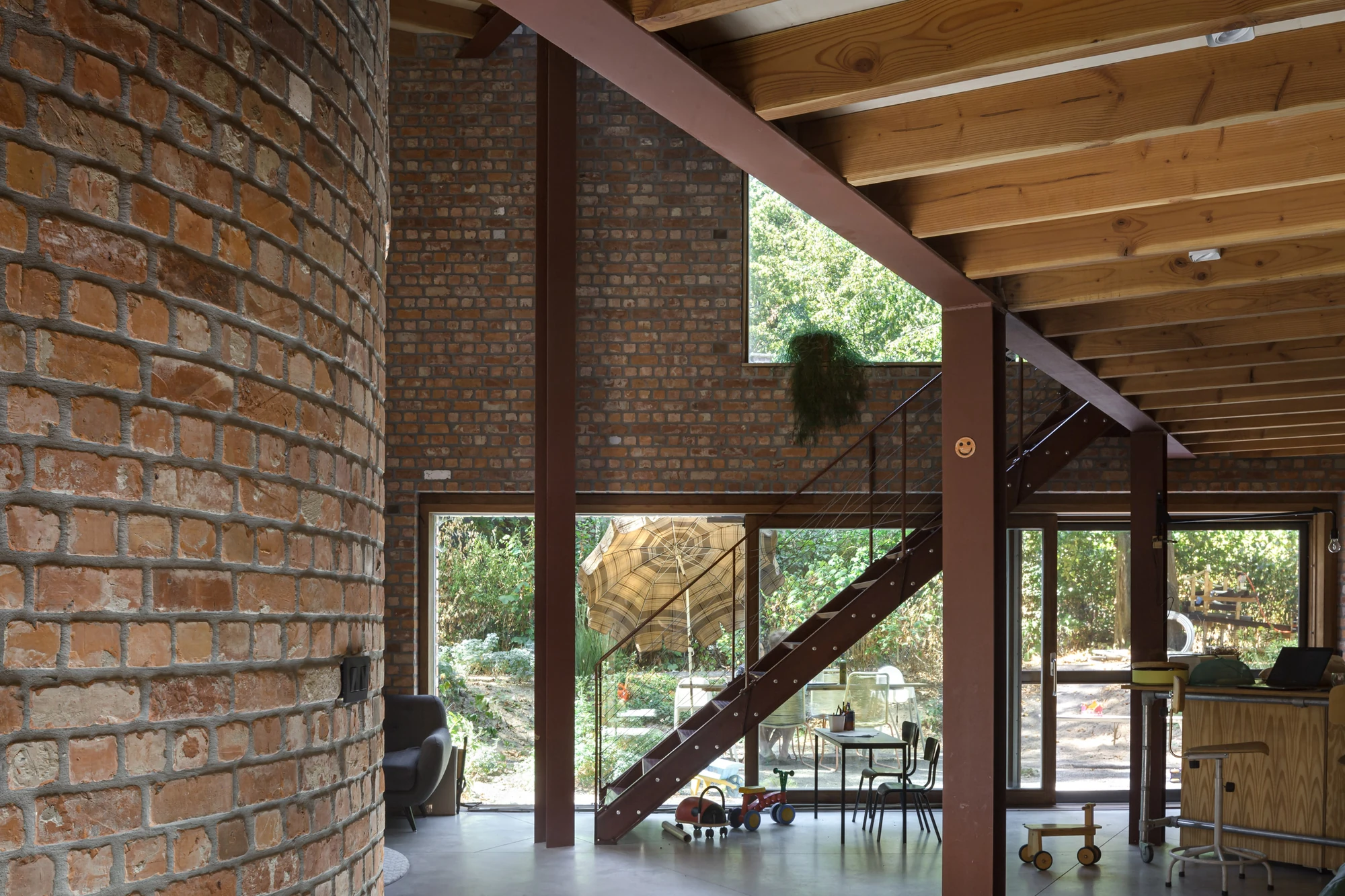 GjG House by BLAF Architecten, photographed by BLAF Architecten — Wide view across the ground floor, brick pier at left, oxblood steel stair rising to the platform level and sliding glazing open onto the garden.