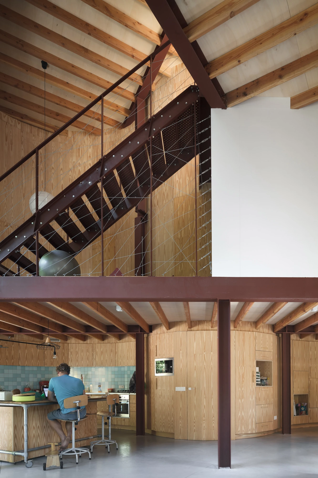 GjG House by BLAF Architecten, photographed by BLAF Architecten — Kitchen below the platform level, oxblood steel stair and cable balustrade above, curved plywood volume behind and a person working at the counter.